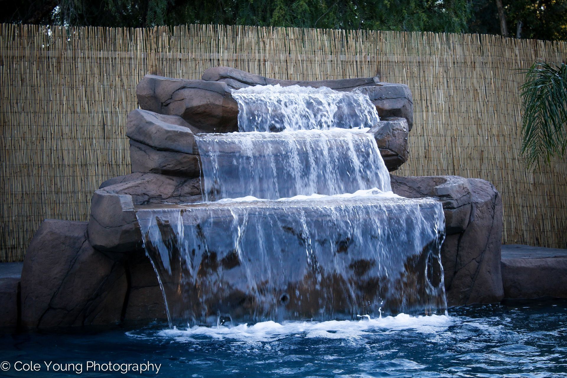 A three-tiered rock waterfall cascading into a pool in front of a woven bamboo fence.