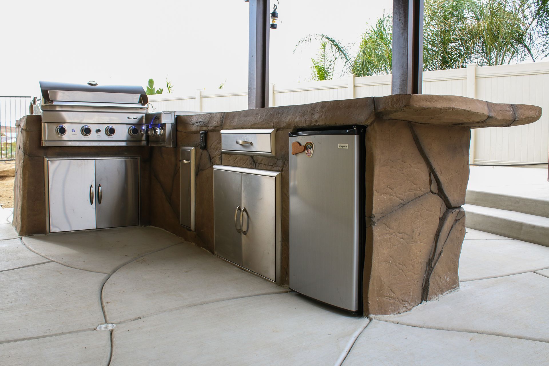Outdoor kitchen island with a built-in grill, stainless steel cabinets, and a mini fridge on a paved patio.
