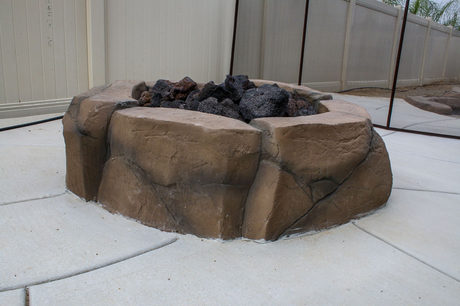 A tan, rock-textured outdoor fire pit filled with dark lava rocks sitting on a concrete patio.