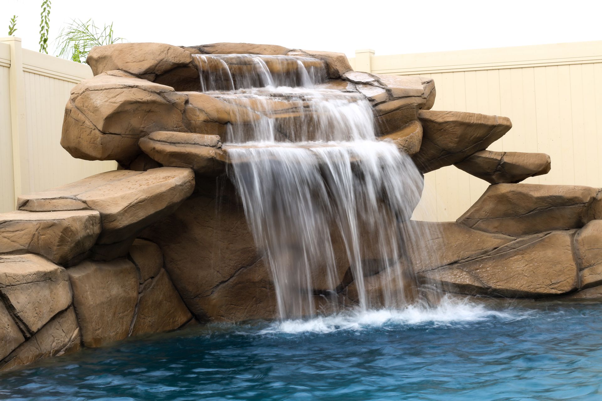 A multi-tiered artificial rock waterfall cascades into a swimming pool against a light-colored fence.