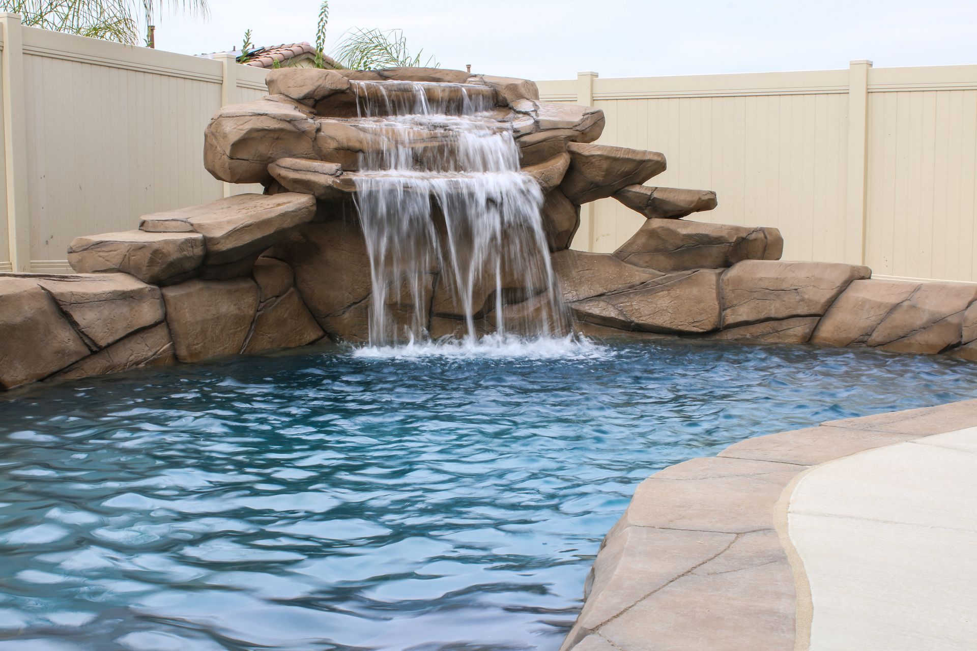 A multi-tiered artificial rock waterfall flowing into a blue swimming pool in a fenced backyard.