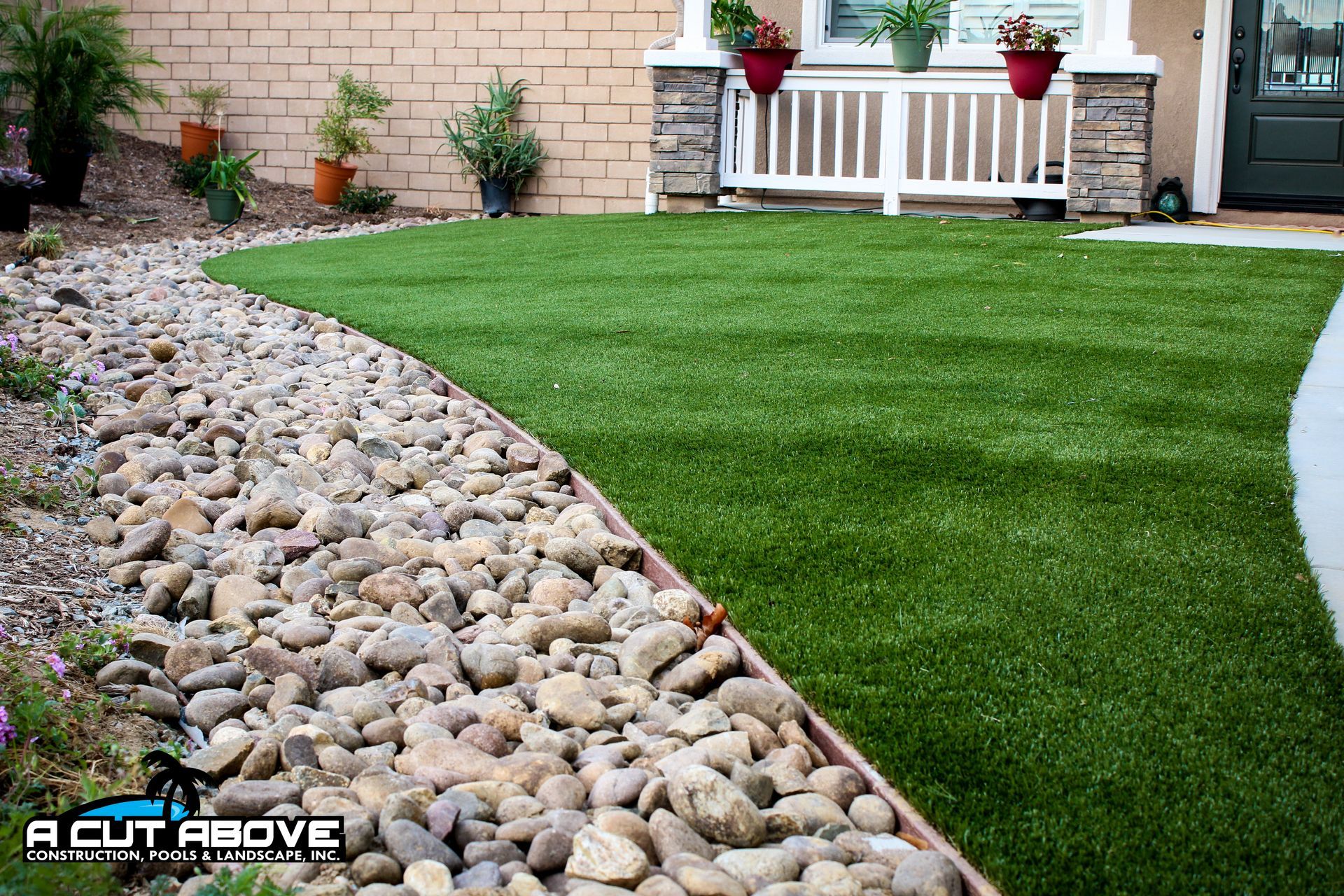 A lush green artificial lawn bordered by decorative river rocks in a residential front yard.
