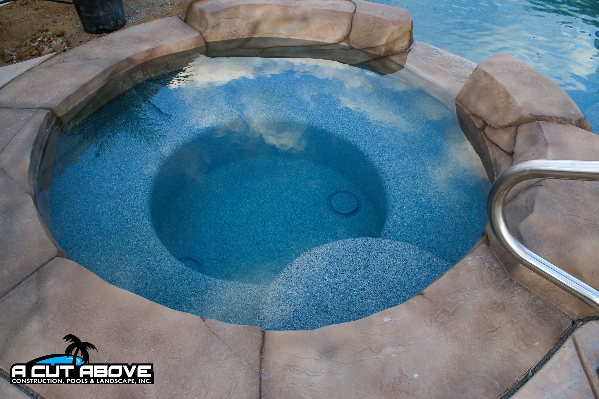 A circular, blue-tiled spa with natural-looking rock coping, partially connected to a larger pool.