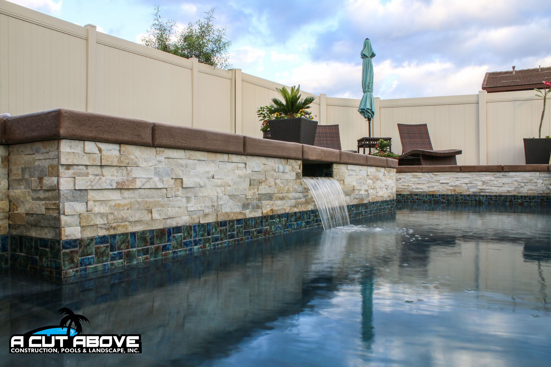 A stone-clad water feature spills into a dark-tiled swimming pool in a backyard with a tan fence and patio furniture.