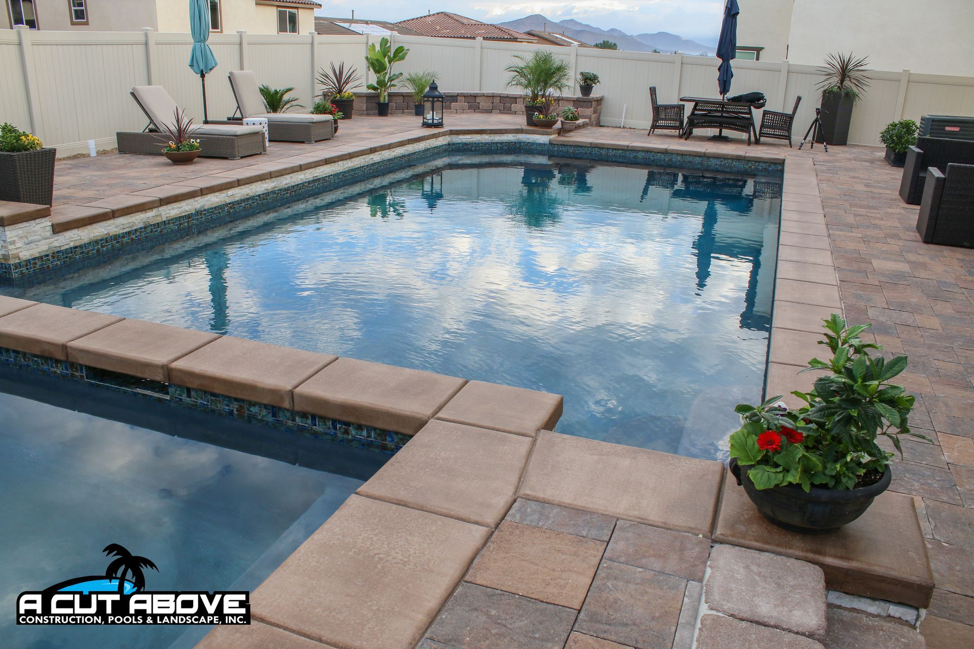 A backyard swimming pool with a spa, stone patio, lounge chairs, and potted plants under a cloudy sky.