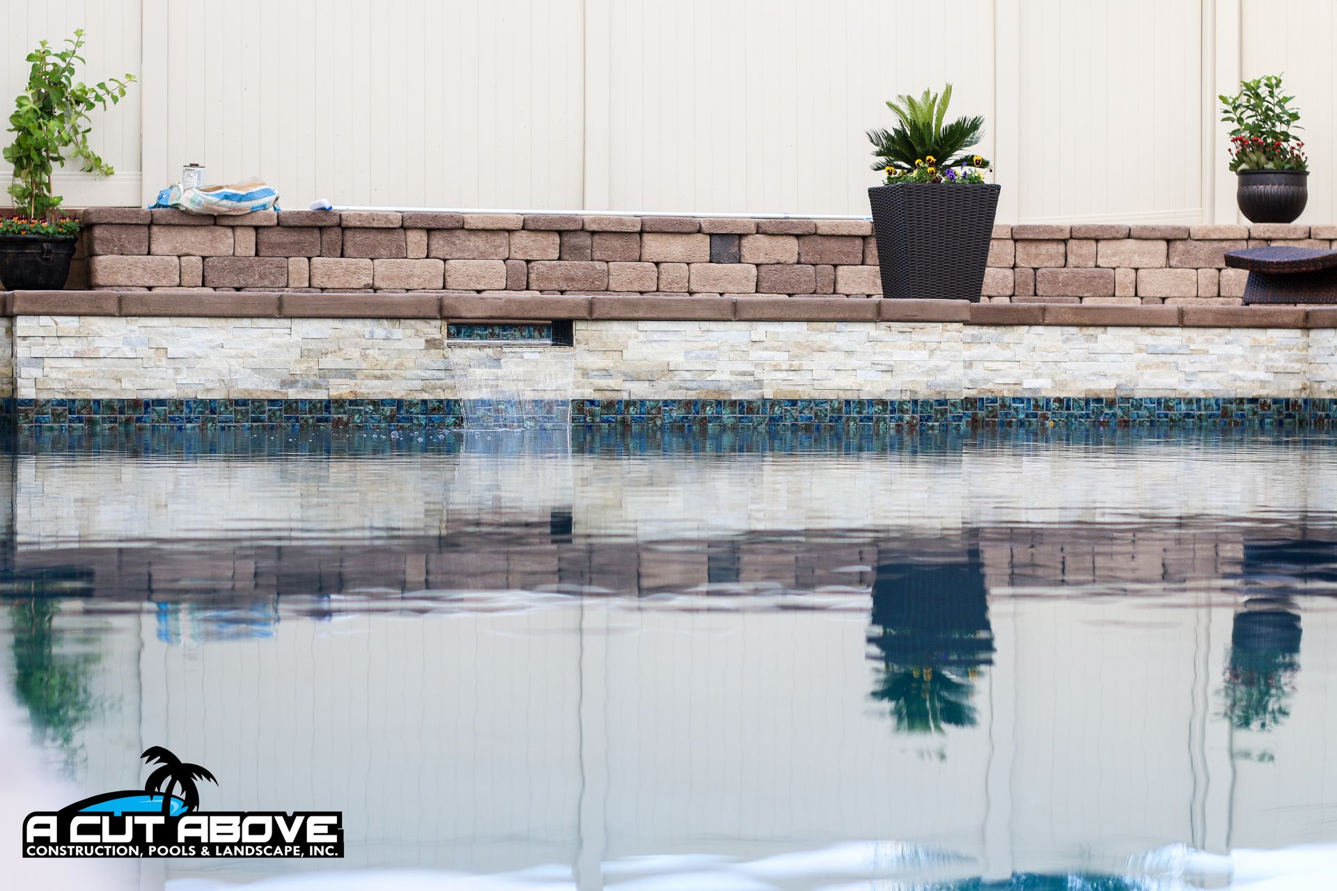 A stone water feature with a fountain stream spills into a blue swimming pool in front of a white privacy fence.