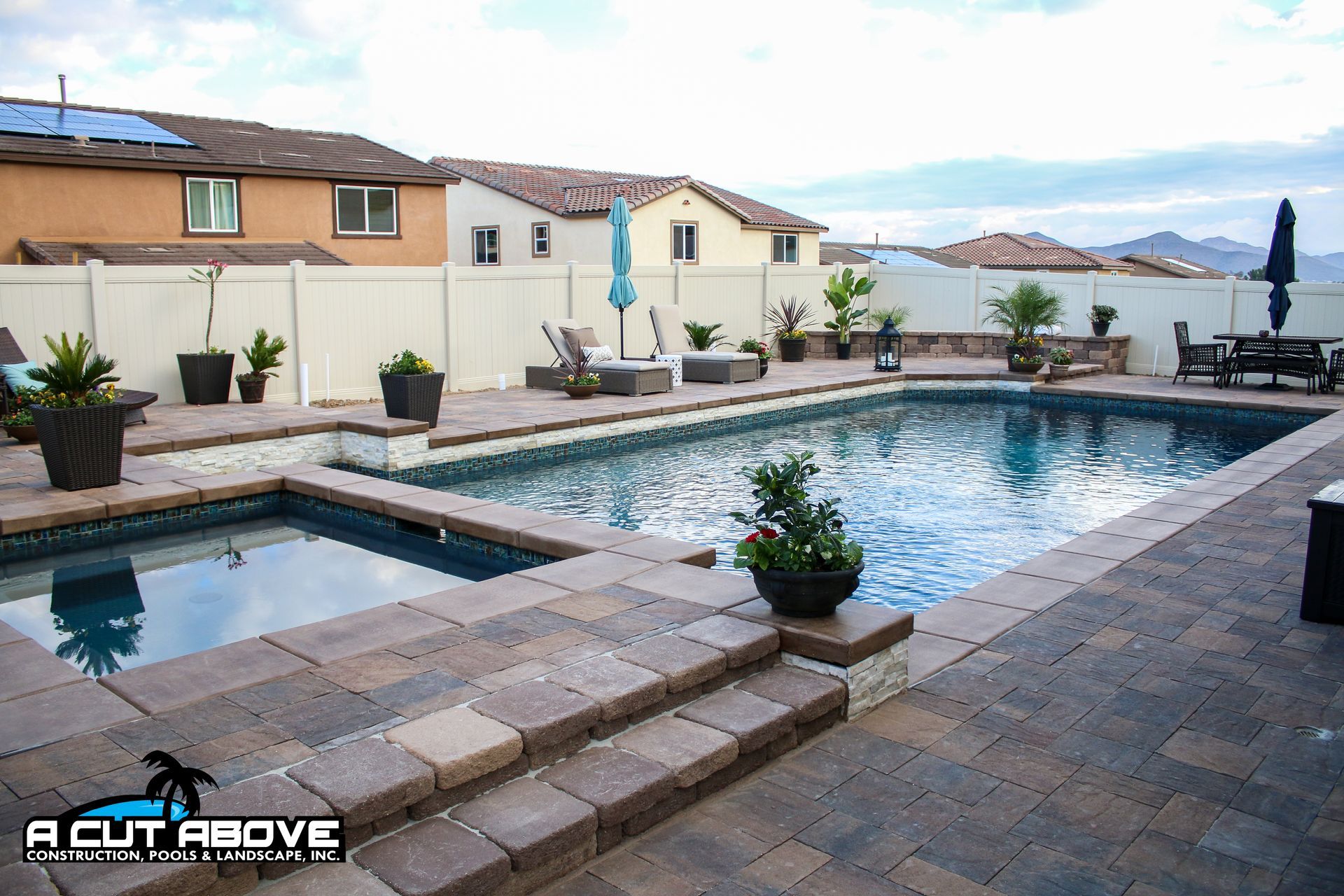 A backyard swimming pool and attached hot tub with paver decking, surrounded by a tan privacy fence and potted plants.