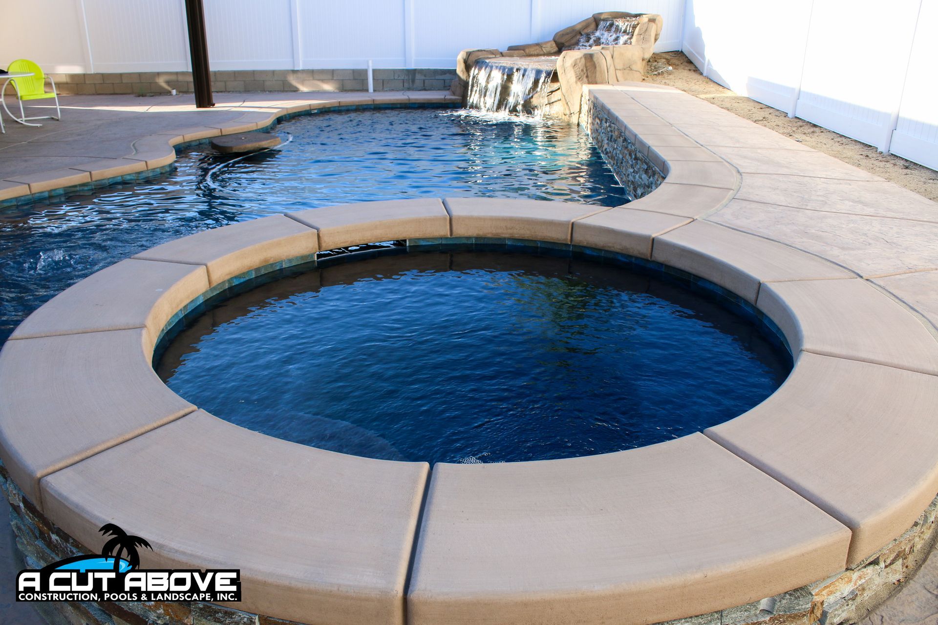 A round hot tub adjacent to a swimming pool with a rock waterfall feature and light-colored concrete decking.