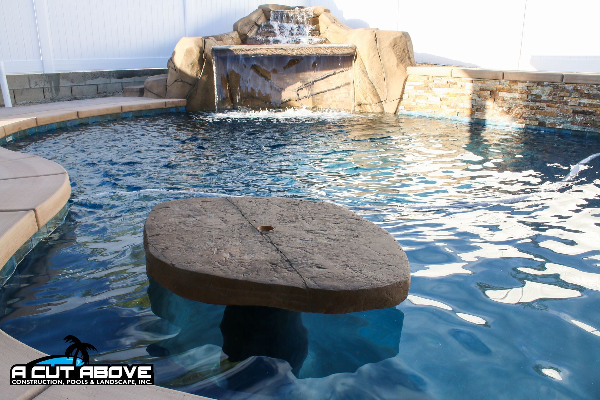 A backyard swimming pool featuring a built-in stone table and a faux-rock waterfall feature with a white privacy fence.