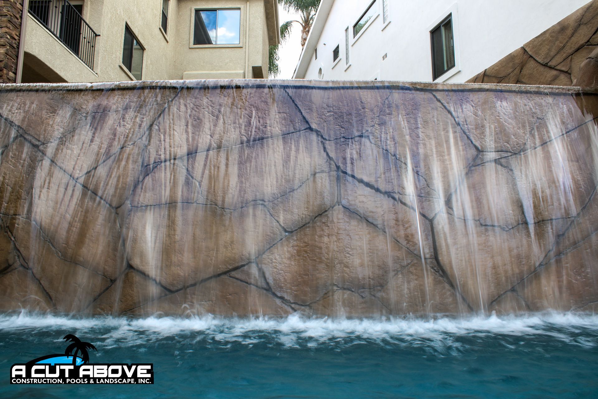 A stone-textured waterfall feature cascades into a swimming pool in front of a multi-story building.