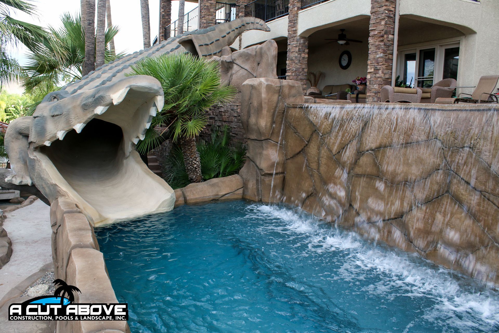 A swimming pool with a large, rock-textured alligator head slide and a stone waterfall feature on a residential patio.
