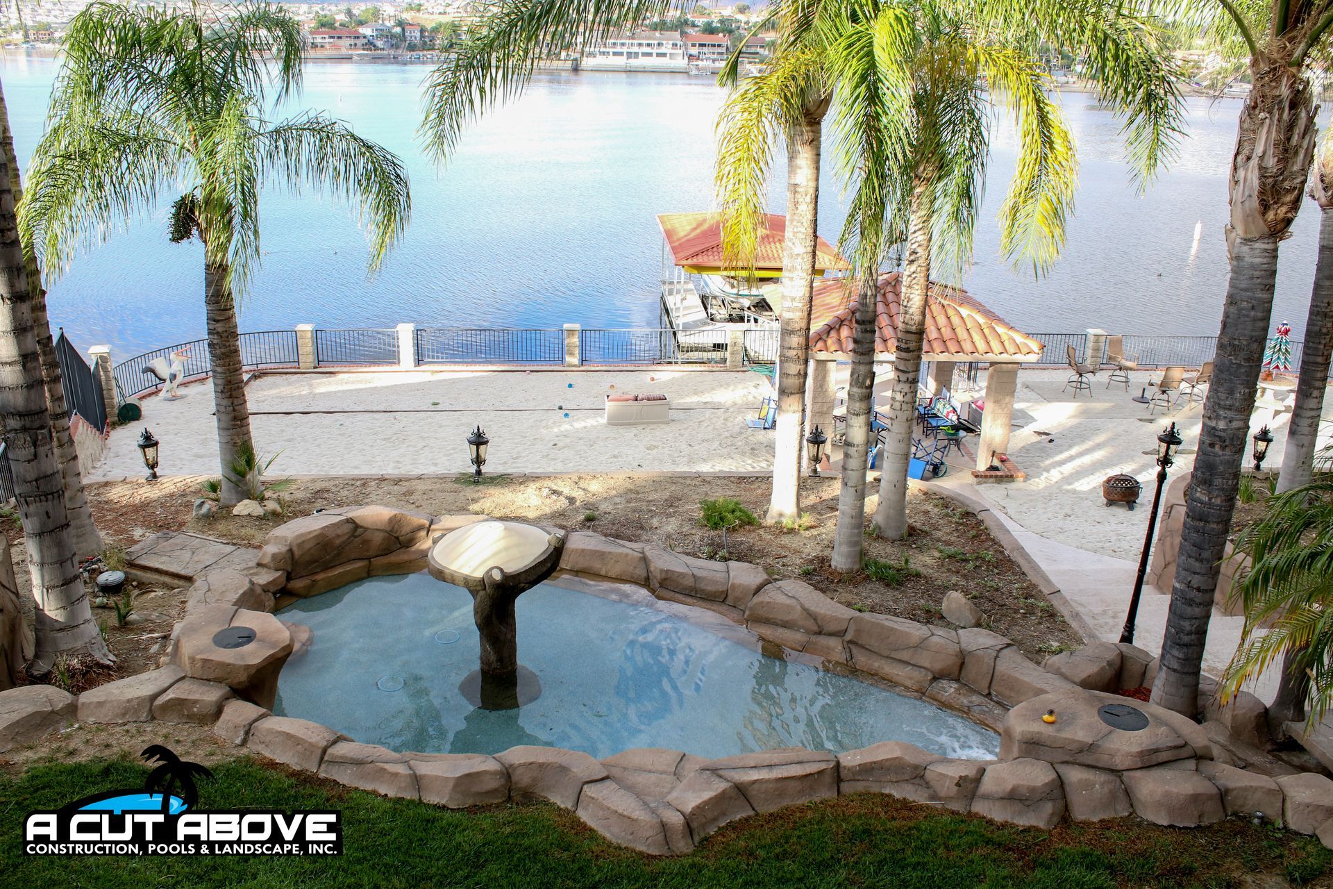 A tiered stone fountain overlooking a swimming pool, palm trees, and a lake with a dock on a sunny day.