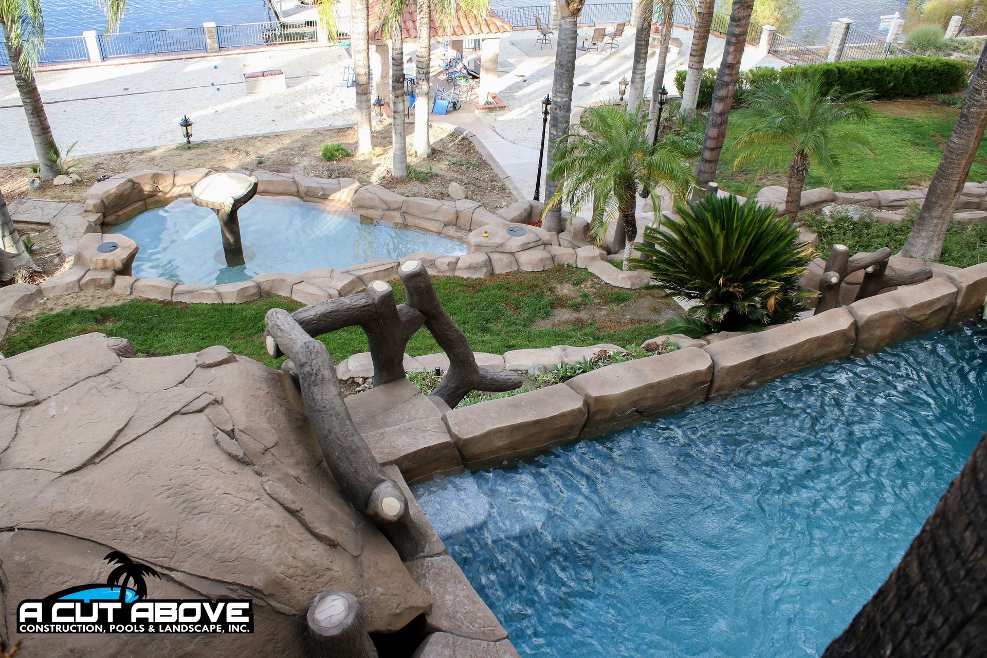 A multi-level pool with faux-rock coping, a stone waterfall, and a small bridge, set in a landscaped hillside yard.