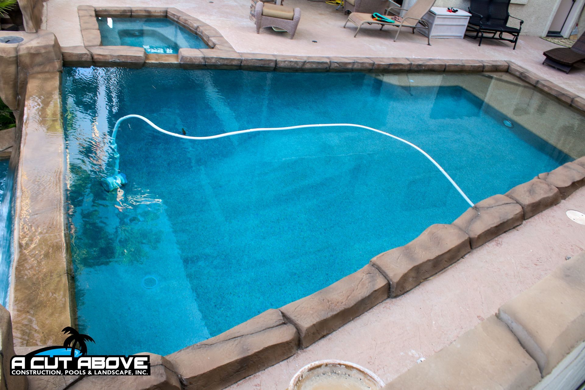 A rectangular backyard swimming pool with a stone coping edge and a connected spa, featuring a pool cleaner hose.