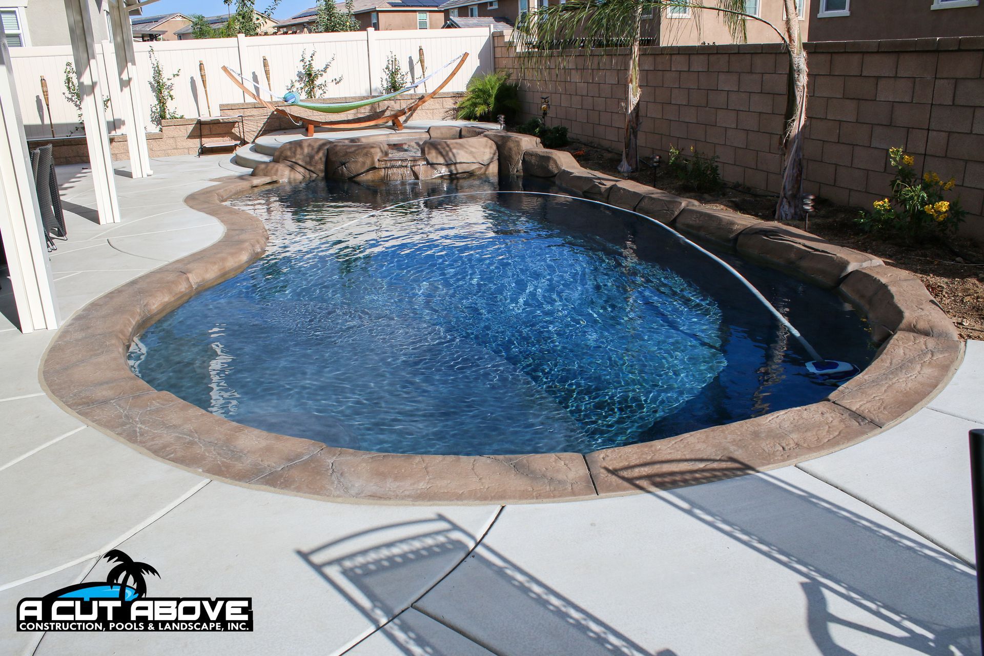 A backyard swimming pool with a rock waterfall feature, concrete decking, and landscaping in a residential setting.