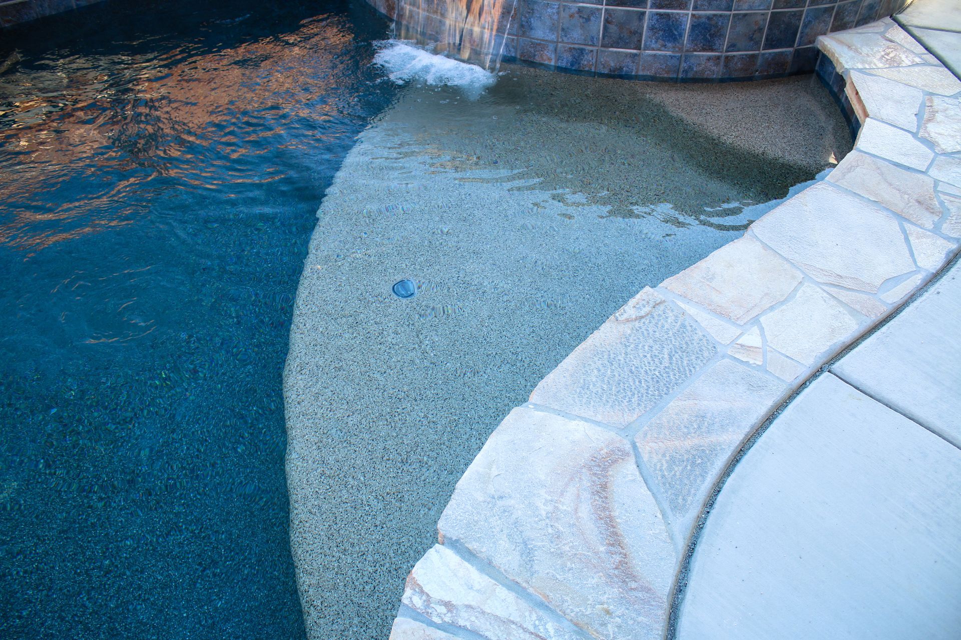 A shallow, sunlit pool entrance with a stone-tiled edge and a waterfall spilling into the deep blue water.
