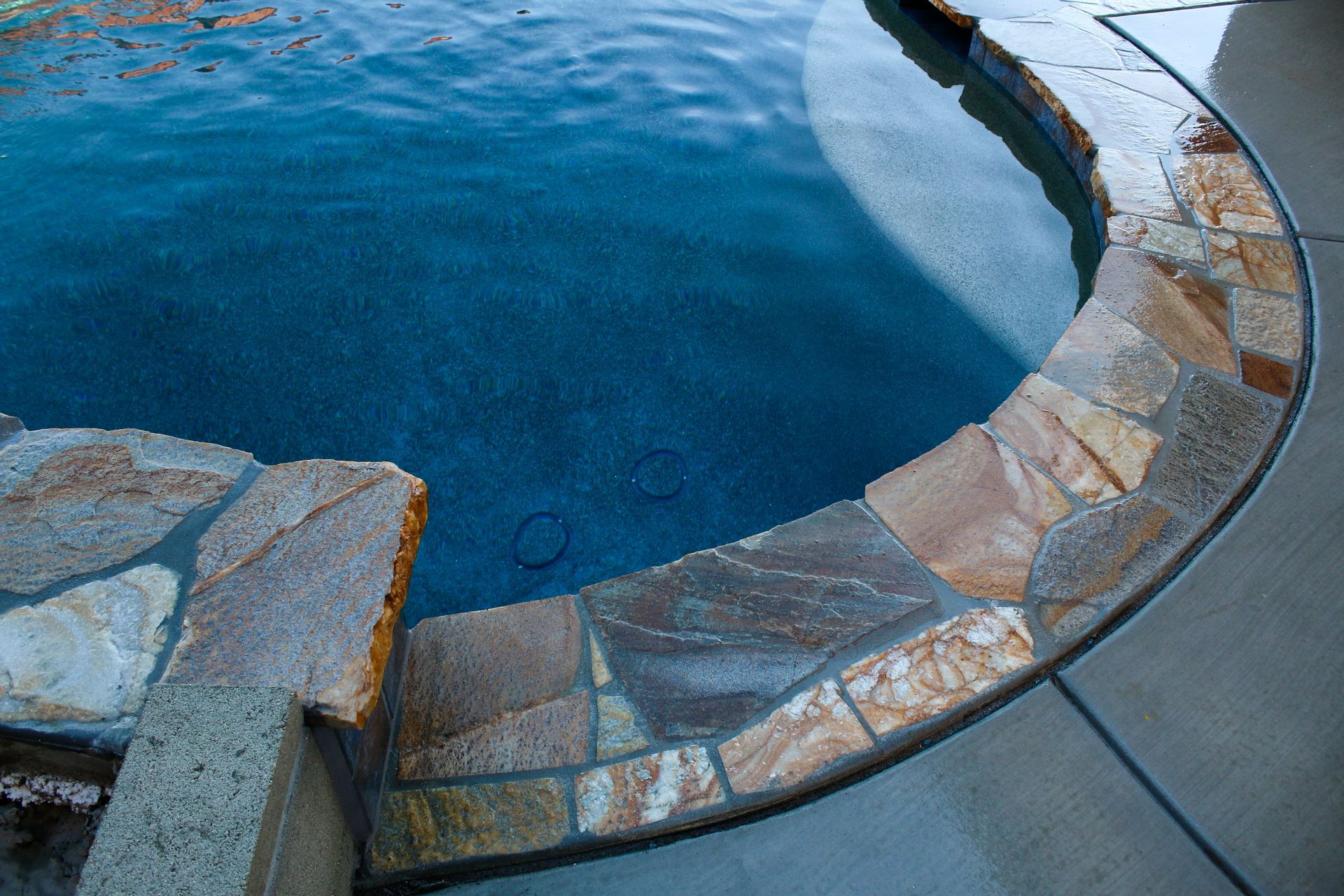 A curved pool edge features textured, multi-colored stone coping stones bordering a deep blue water surface.