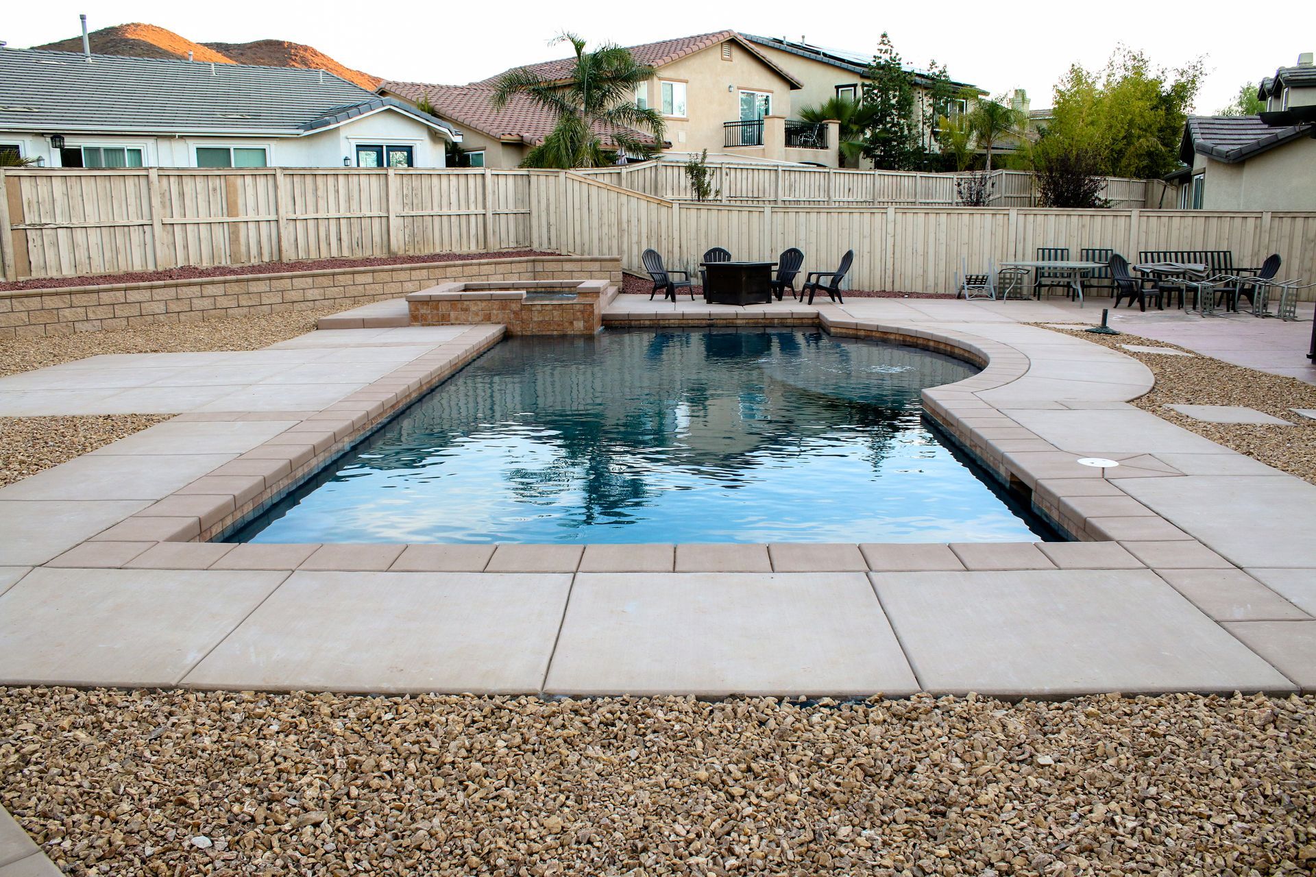 Backyard swimming pool with a stone coping, surrounding concrete patio, and gravel landscaping in a residential area.
