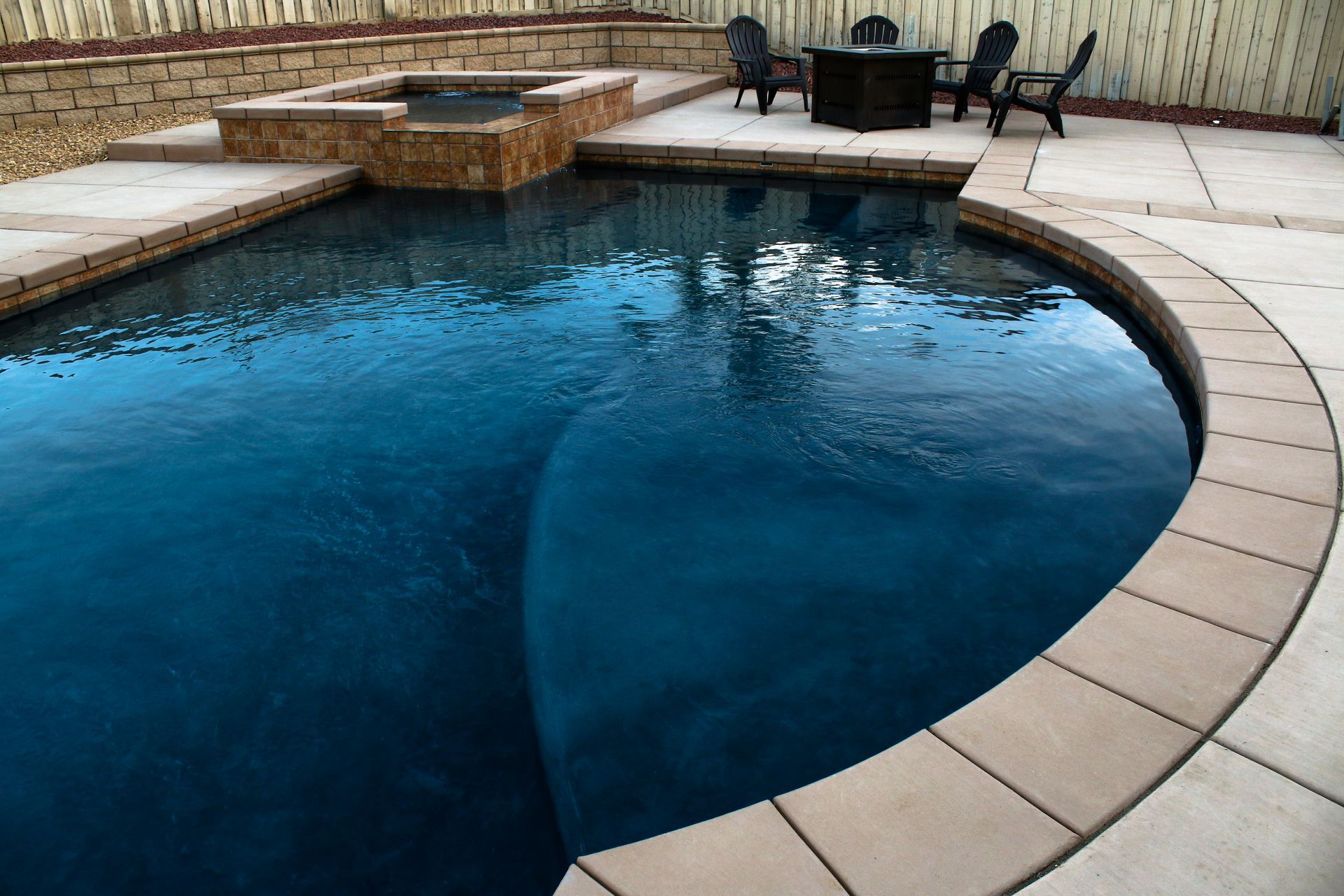 A dark blue swimming pool with curved steps and a stone-trimmed hot tub on a patio with outdoor furniture.