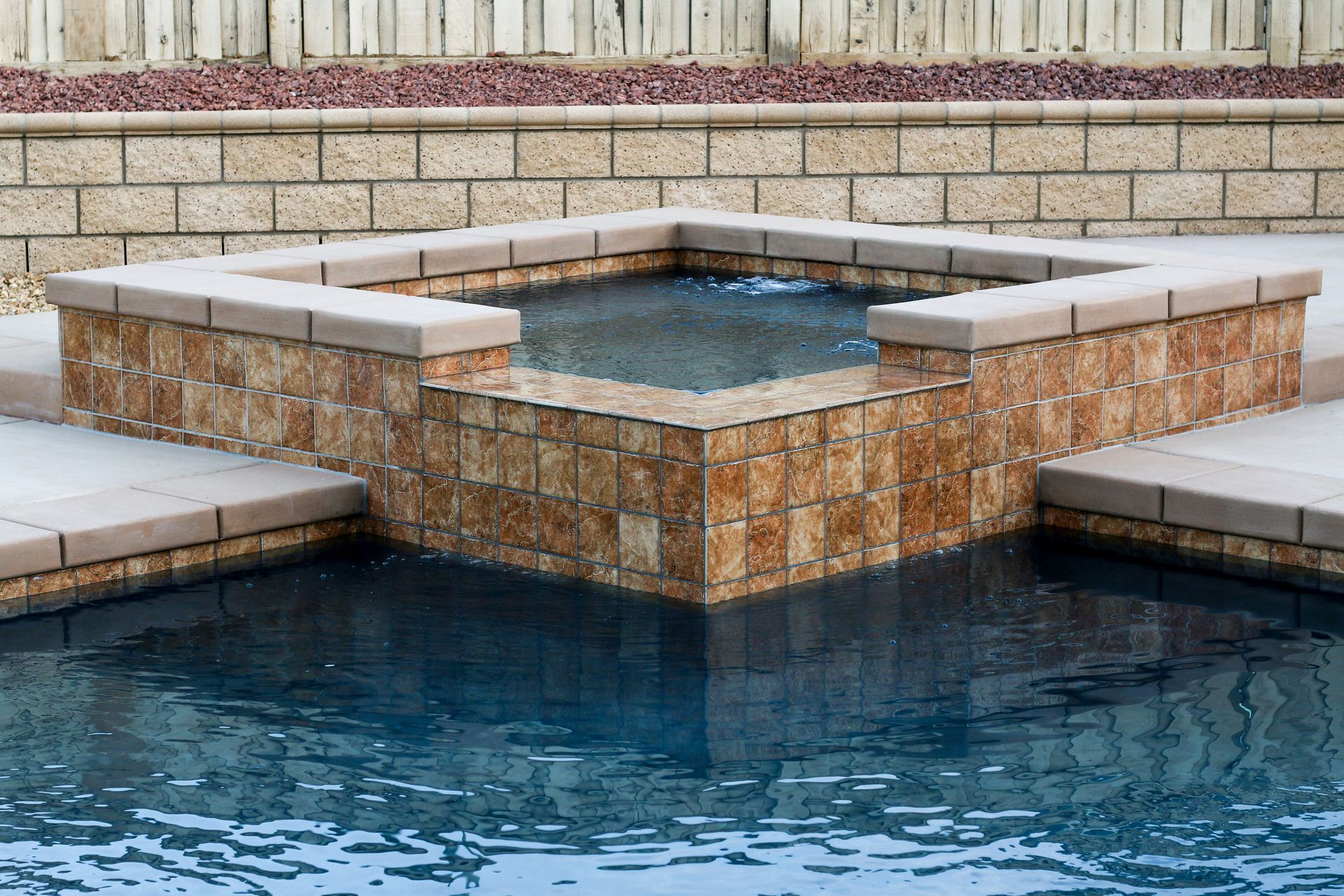 An elevated square spa made of tan textured stone blocks sits directly next to a dark blue swimming pool.