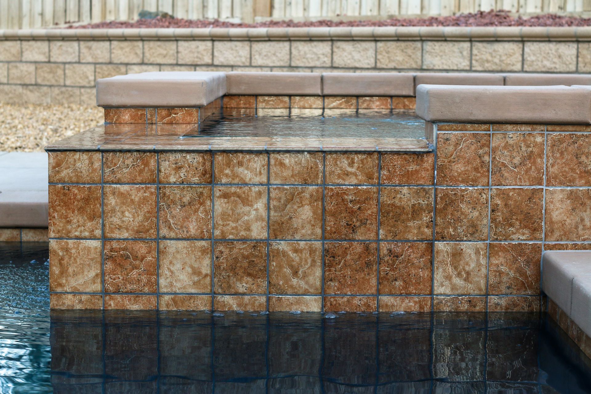 A square spa feature tiled with textured, mottled brown stone, partially submerged in a pool with a stone retaining wall.