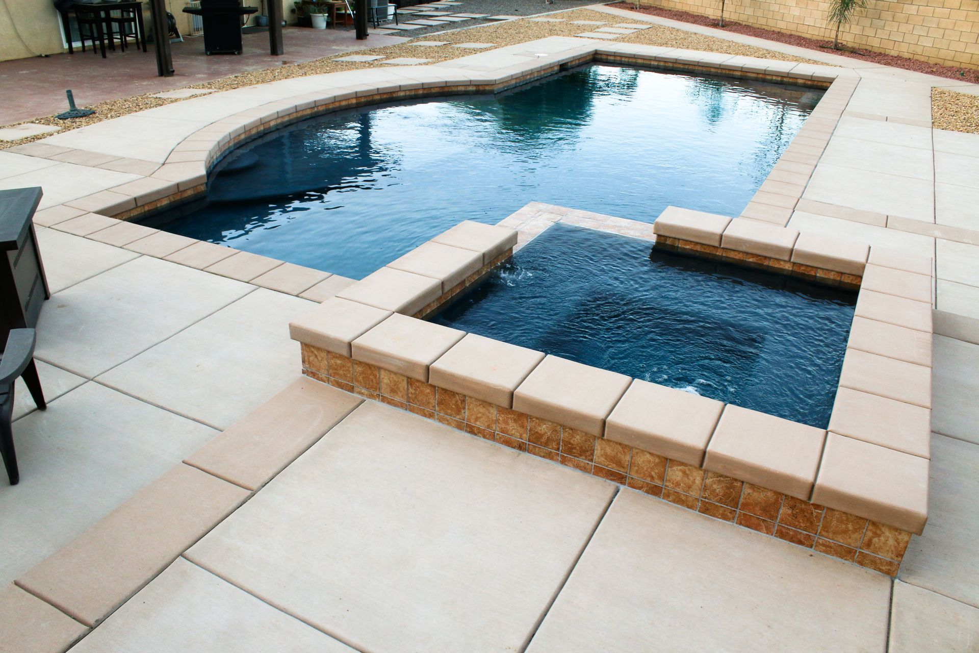 A rectangular swimming pool with a connected, raised square hot tub on a concrete patio in a backyard.