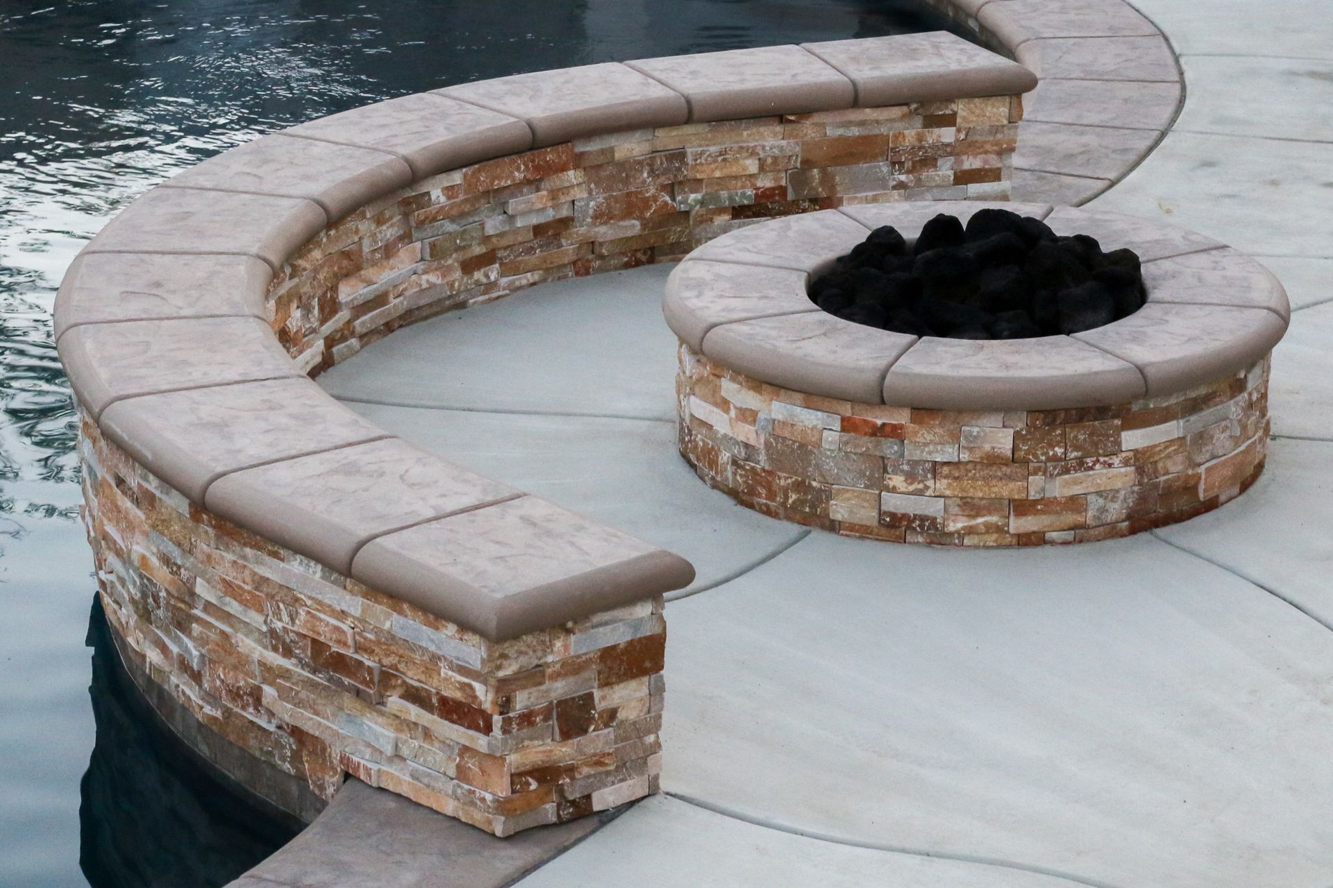 A curved stone seat and a circular stone fire pit with black rocks on a light concrete patio next to a swimming pool.
