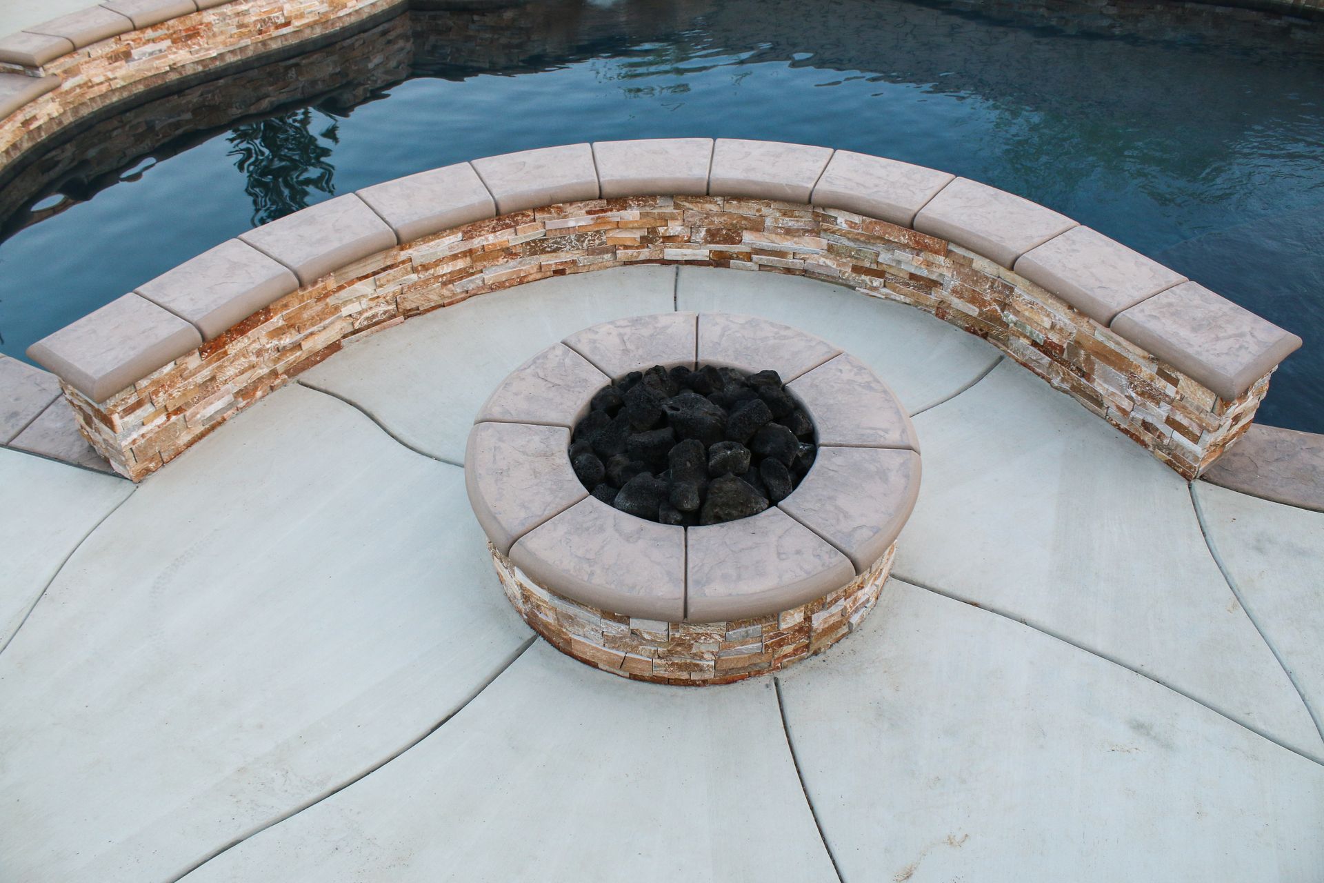 A circular stone fire pit sits on a concrete patio next to a curved stone bench and a swimming pool.
