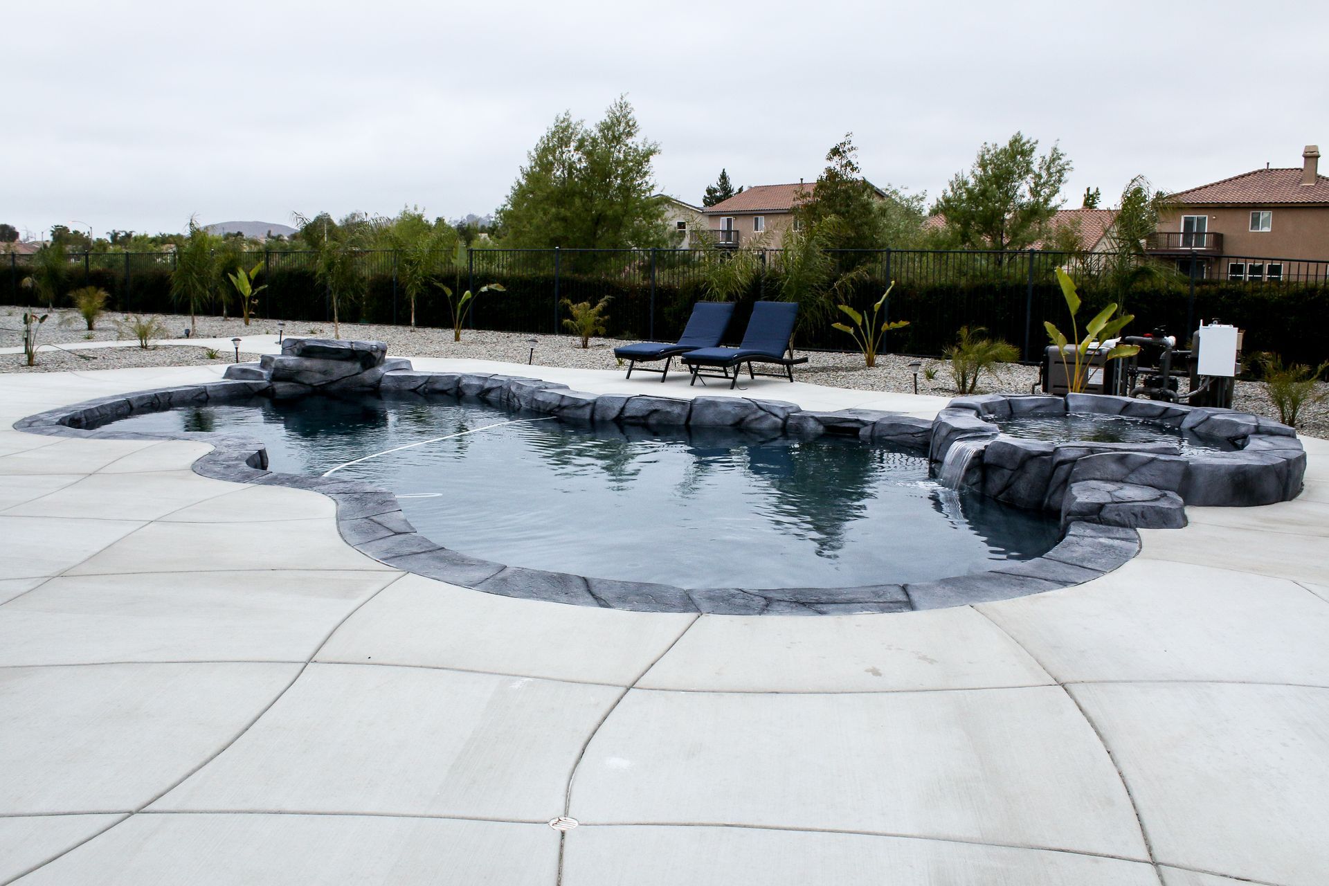A dark-water swimming pool with stone rock features and waterfalls, surrounded by a light concrete deck and landscape.