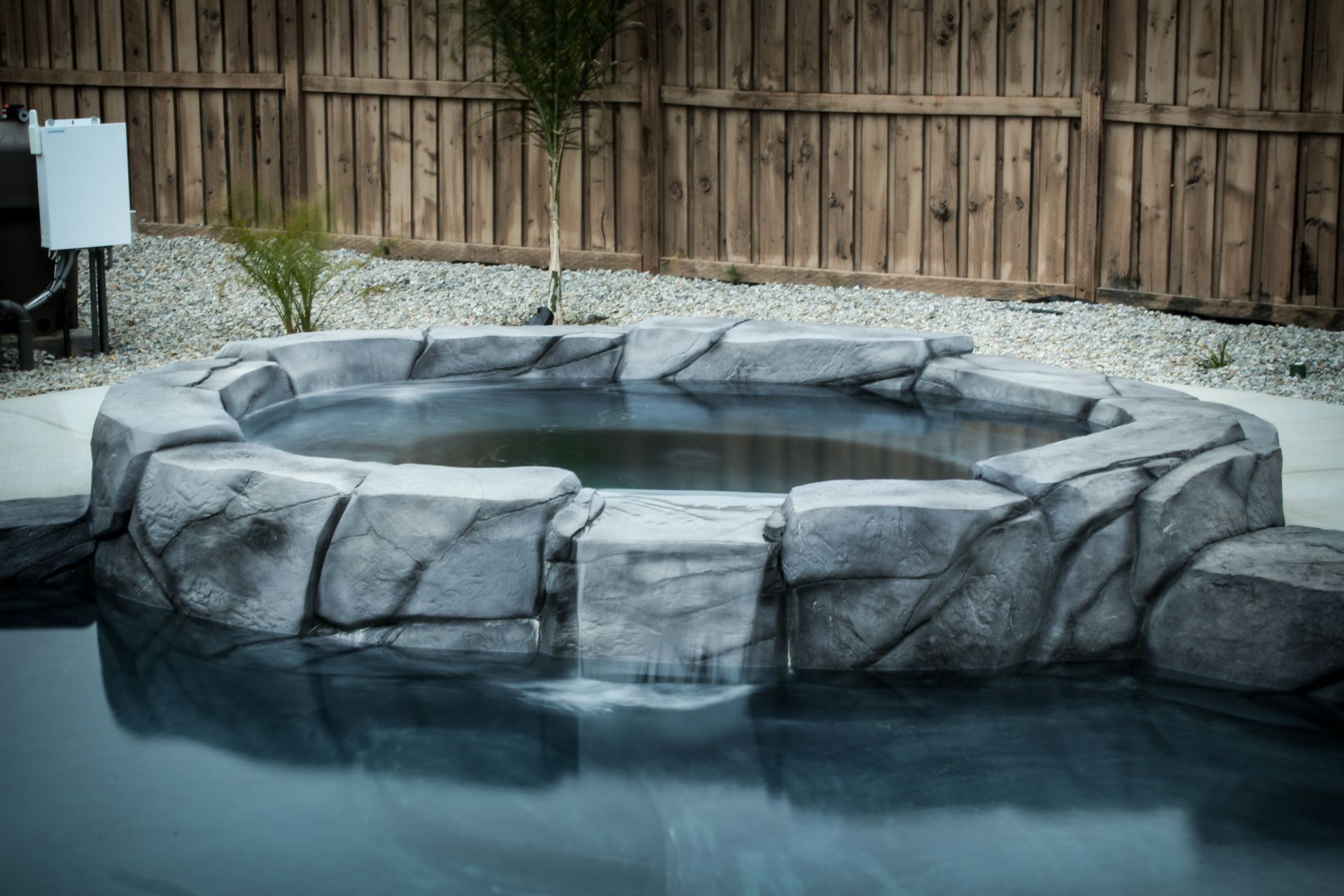 A raised, faux-stone hot tub sits beside a pool in a backyard with a wooden fence and gravel landscaping.
