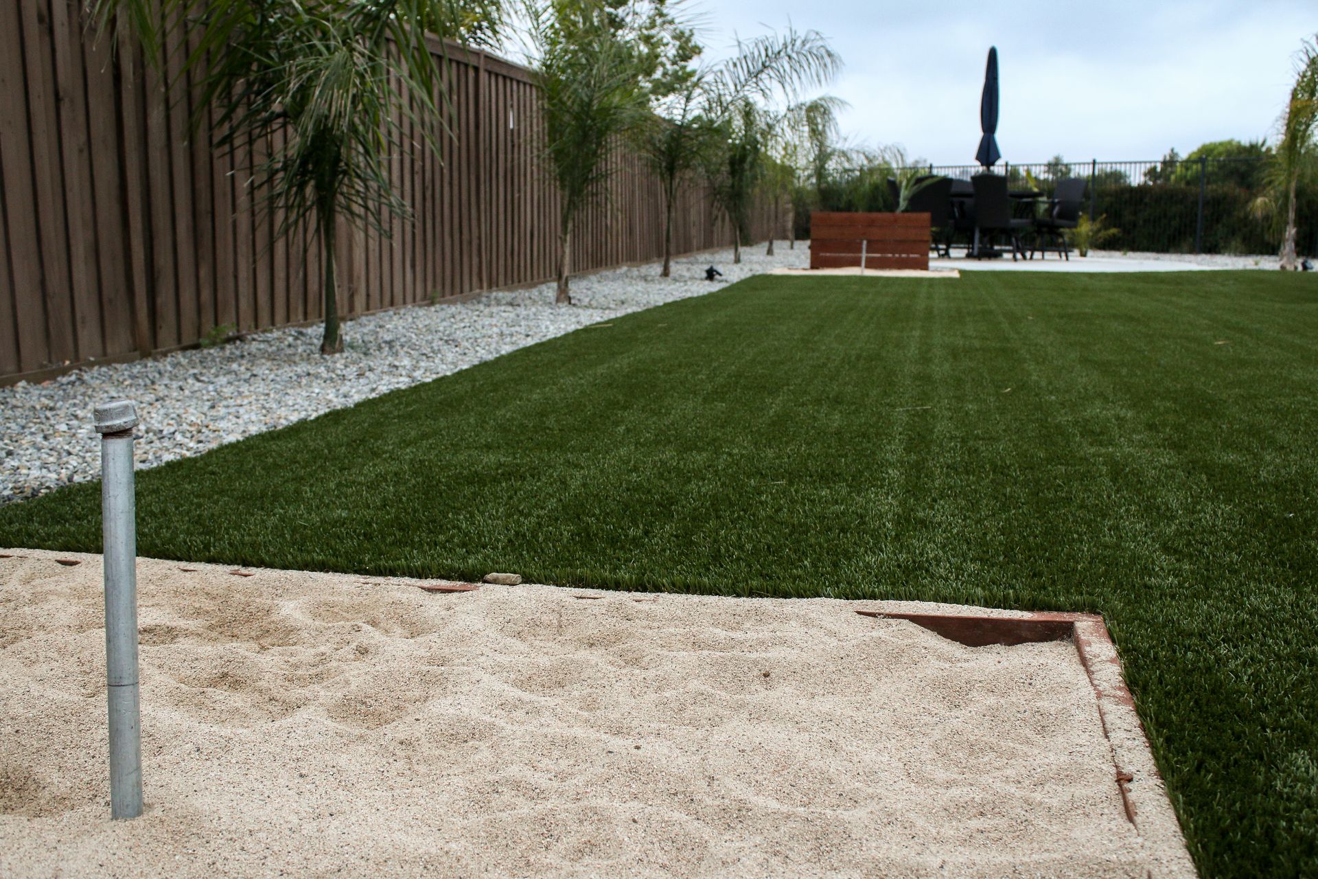 A sand pit foregrounds a vibrant green lawn with a white rock border and tall wooden fence.