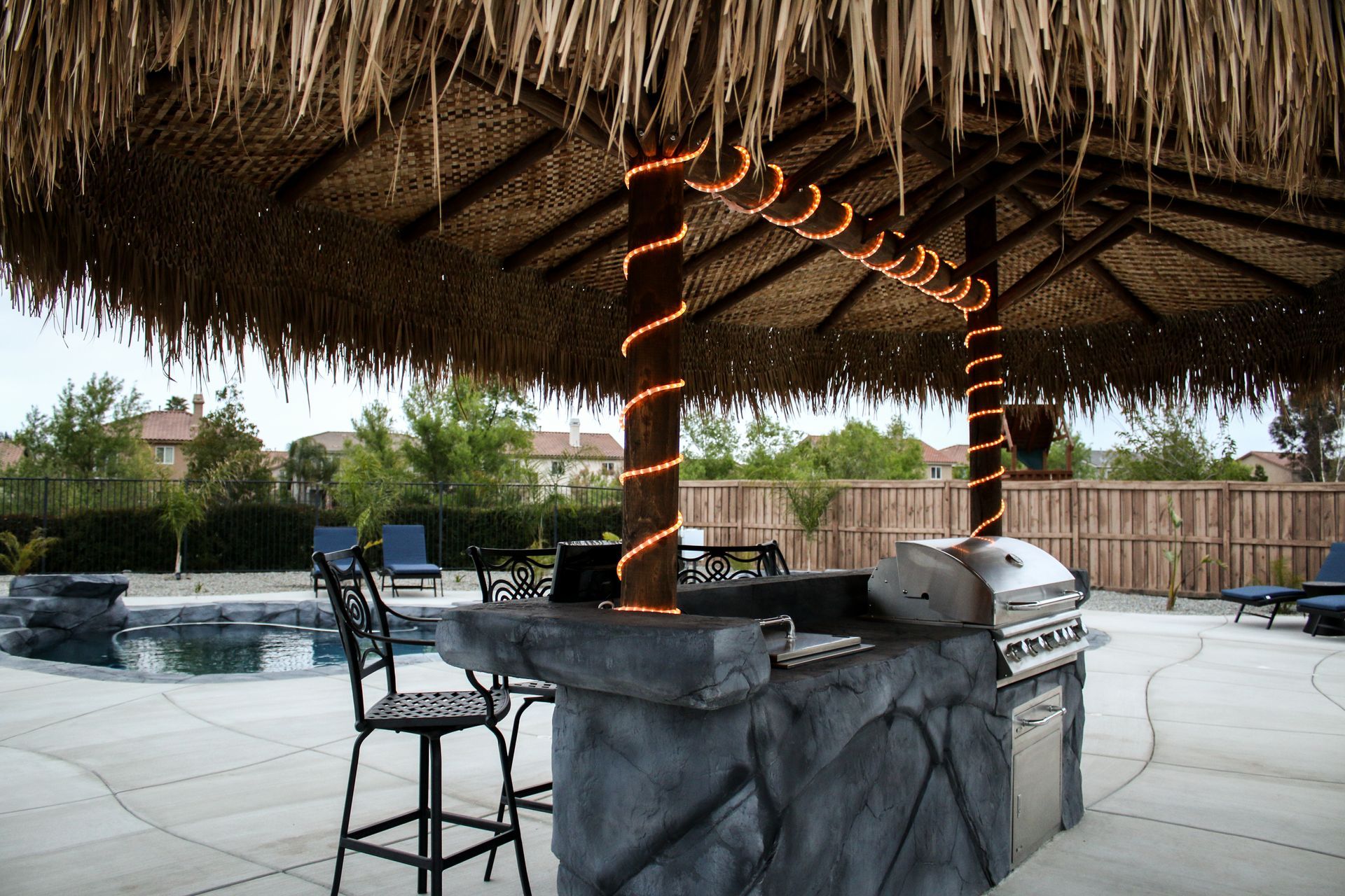 Outdoor tiki-style bar with a thatched roof, glowing string lights, and a stone-look grill station by a backyard pool.