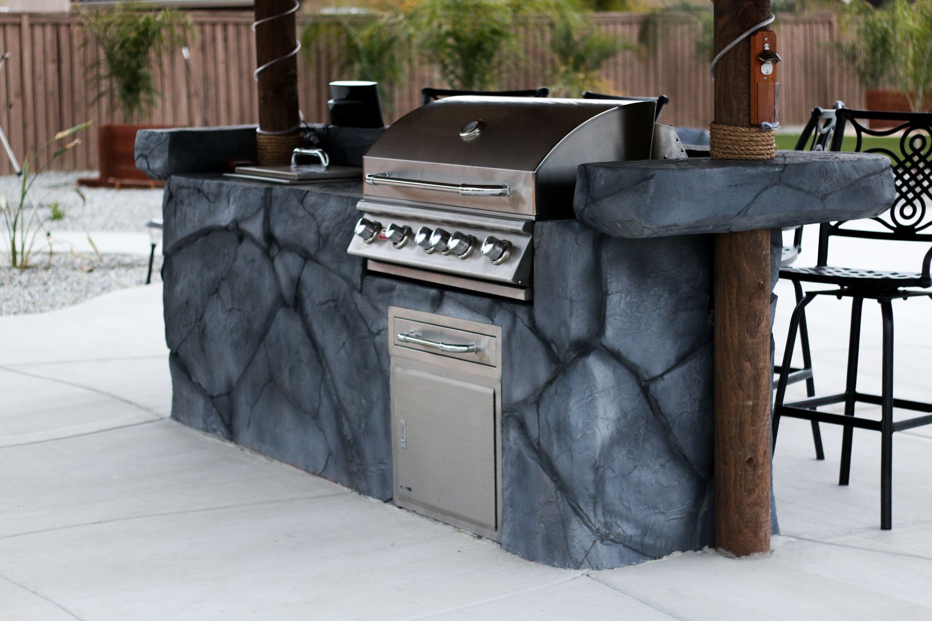 An outdoor kitchen with a stainless steel grill, stone-textured base, and a wooden pillar on a concrete patio.