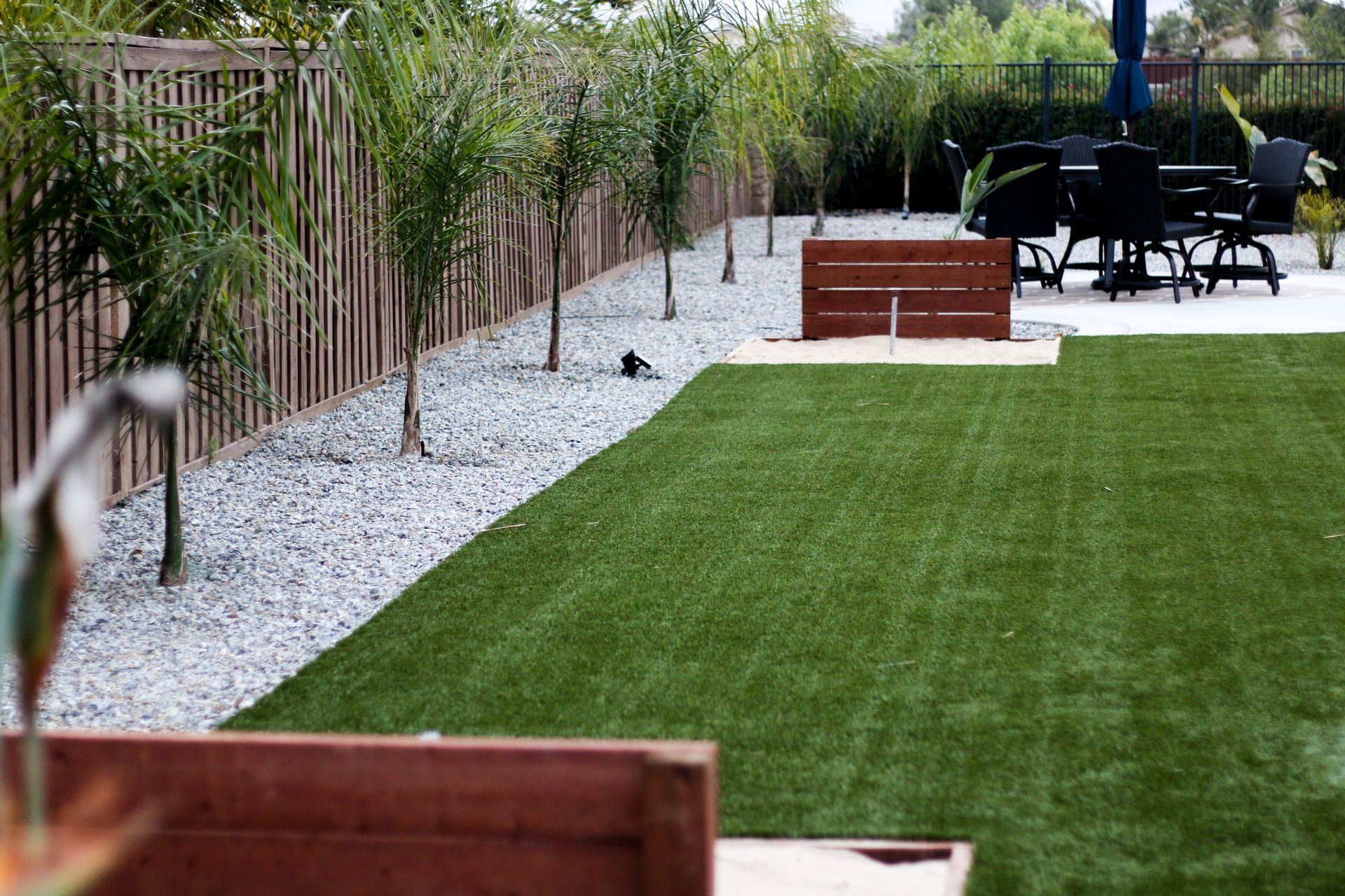 A backyard with a green artificial turf lawn, a gravel border with young palm trees, a wooden fence, and patio furniture.