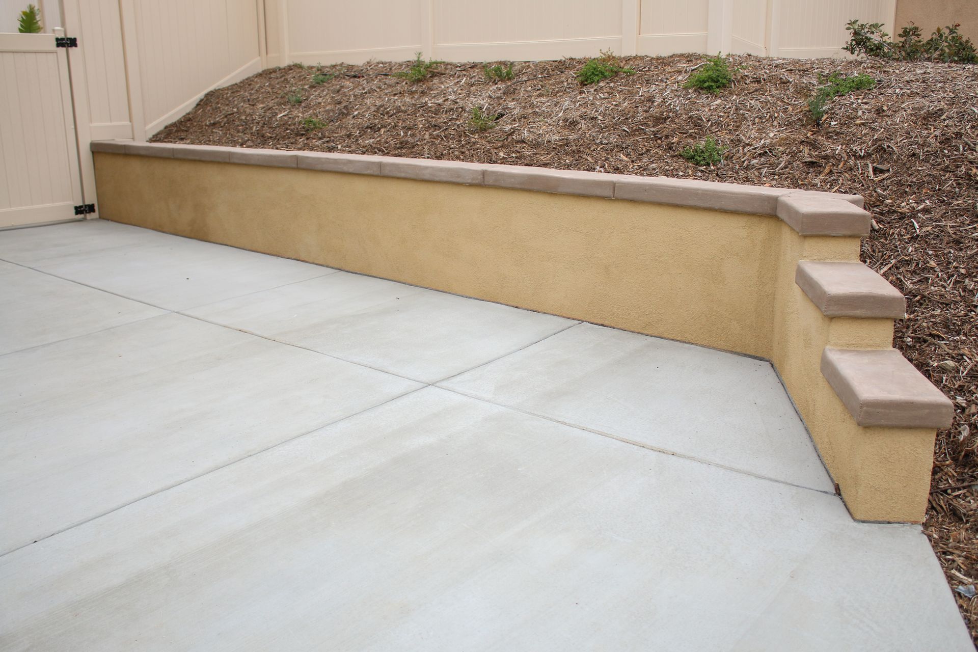 A beige stucco retaining wall with a tiered edge sits beside a concrete patio, with a mulched slope in the background.