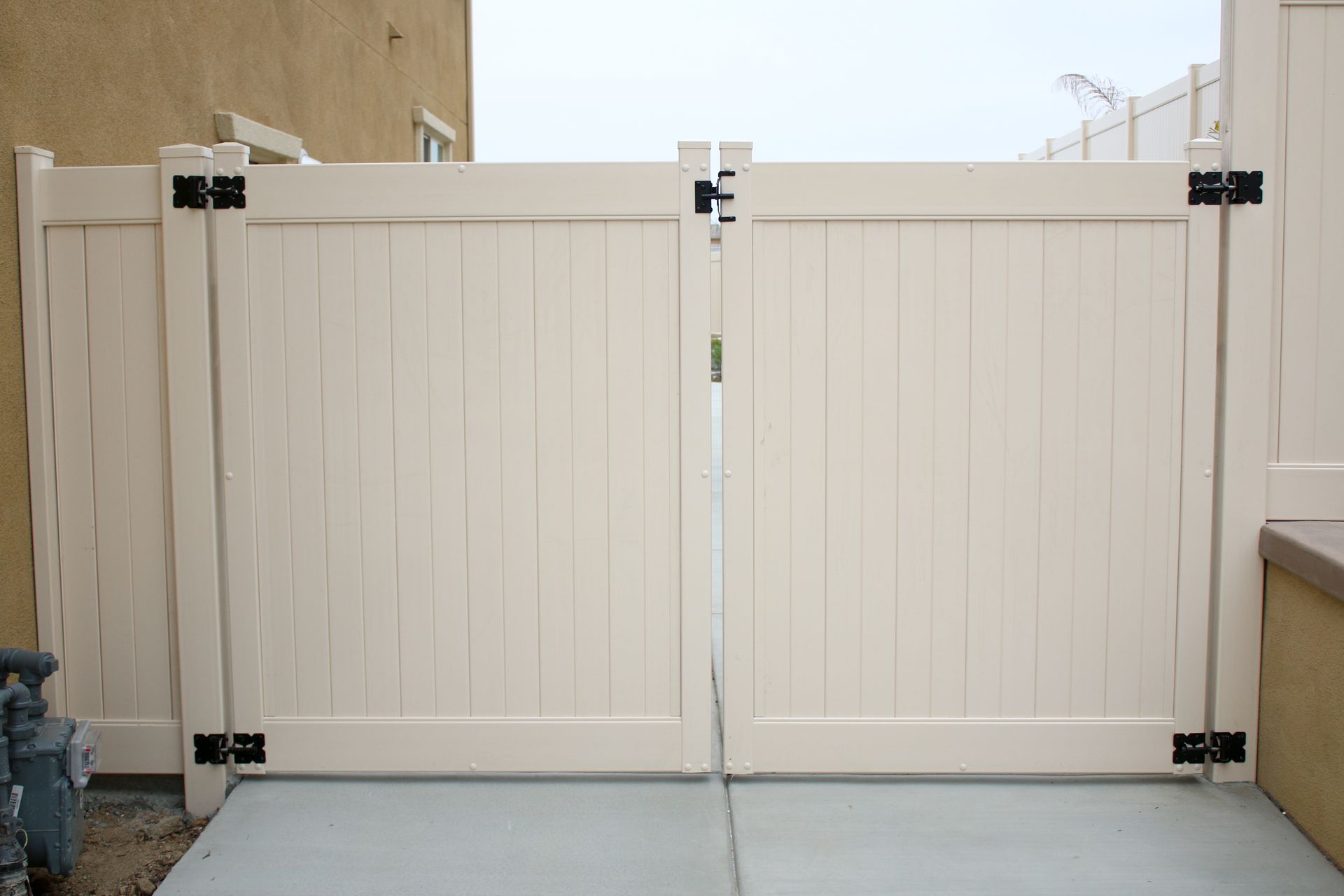 A double vinyl gate with vertical slats and black hinges in a light tan color, situated on a concrete patio.