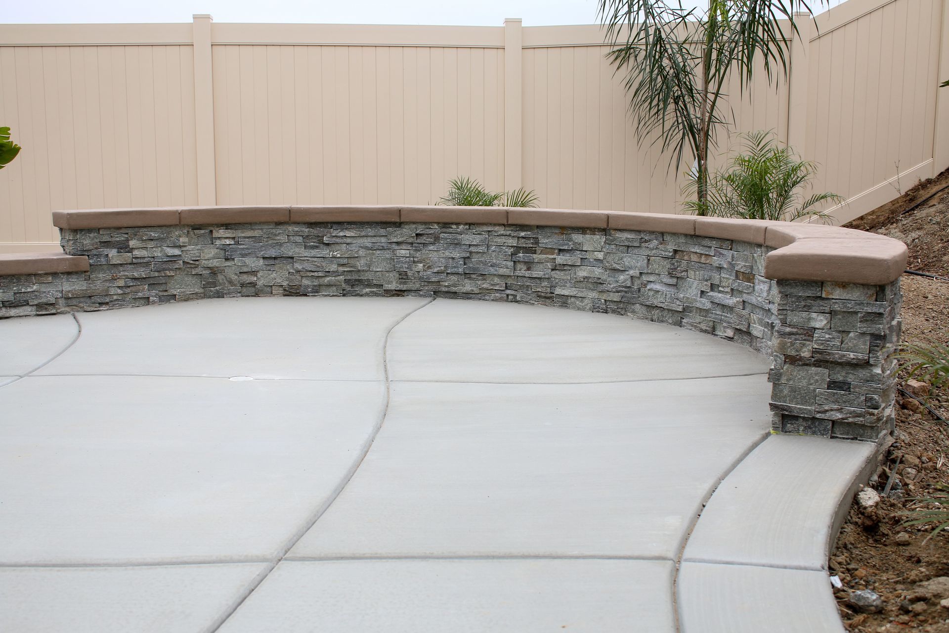 A semi-circular, stacked-stone low wall with a smooth, tan cap sits on a curved concrete patio against a beige fence.