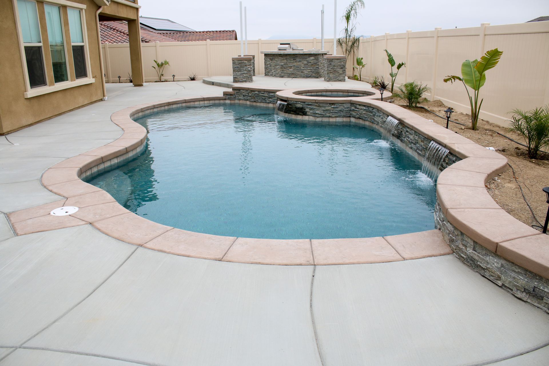 A backyard pool with a built-in spa, stone water features, and a concrete patio next to a tan house and privacy fence.