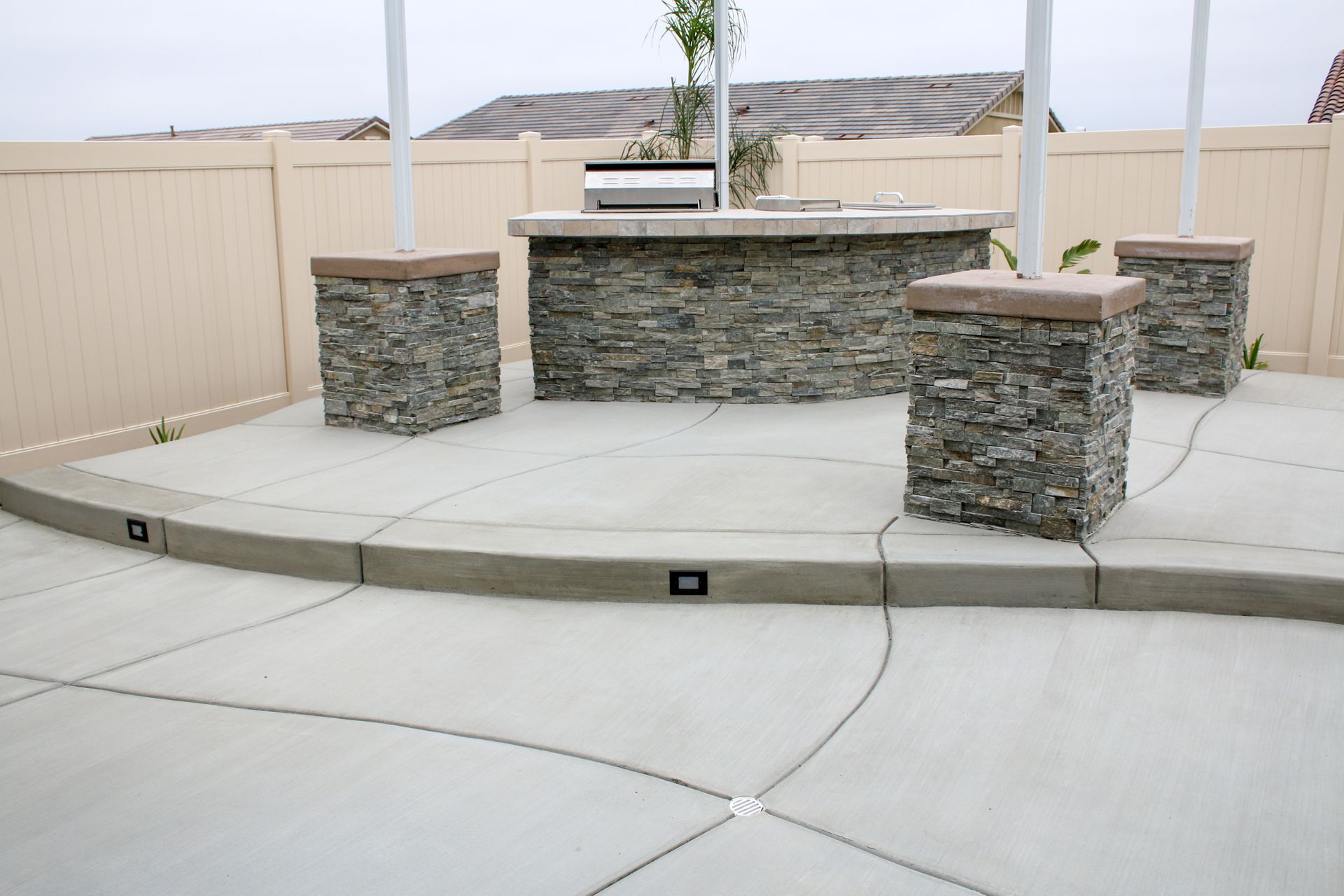An outdoor patio with a stone-accented bar, grill, and decorative concrete floor, enclosed by a tan vinyl privacy fence.