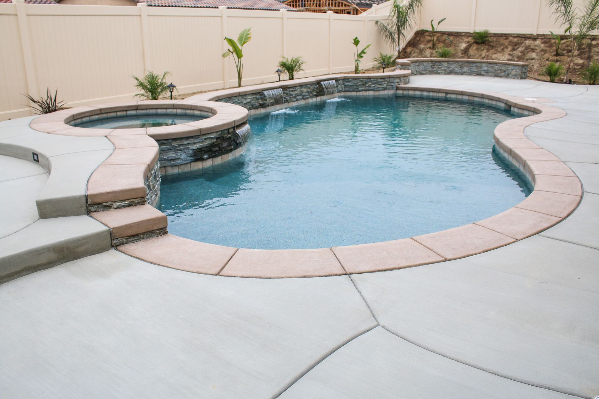 A swimming pool and attached hot tub with tan stone coping, set in a backyard patio with a beige perimeter fence.