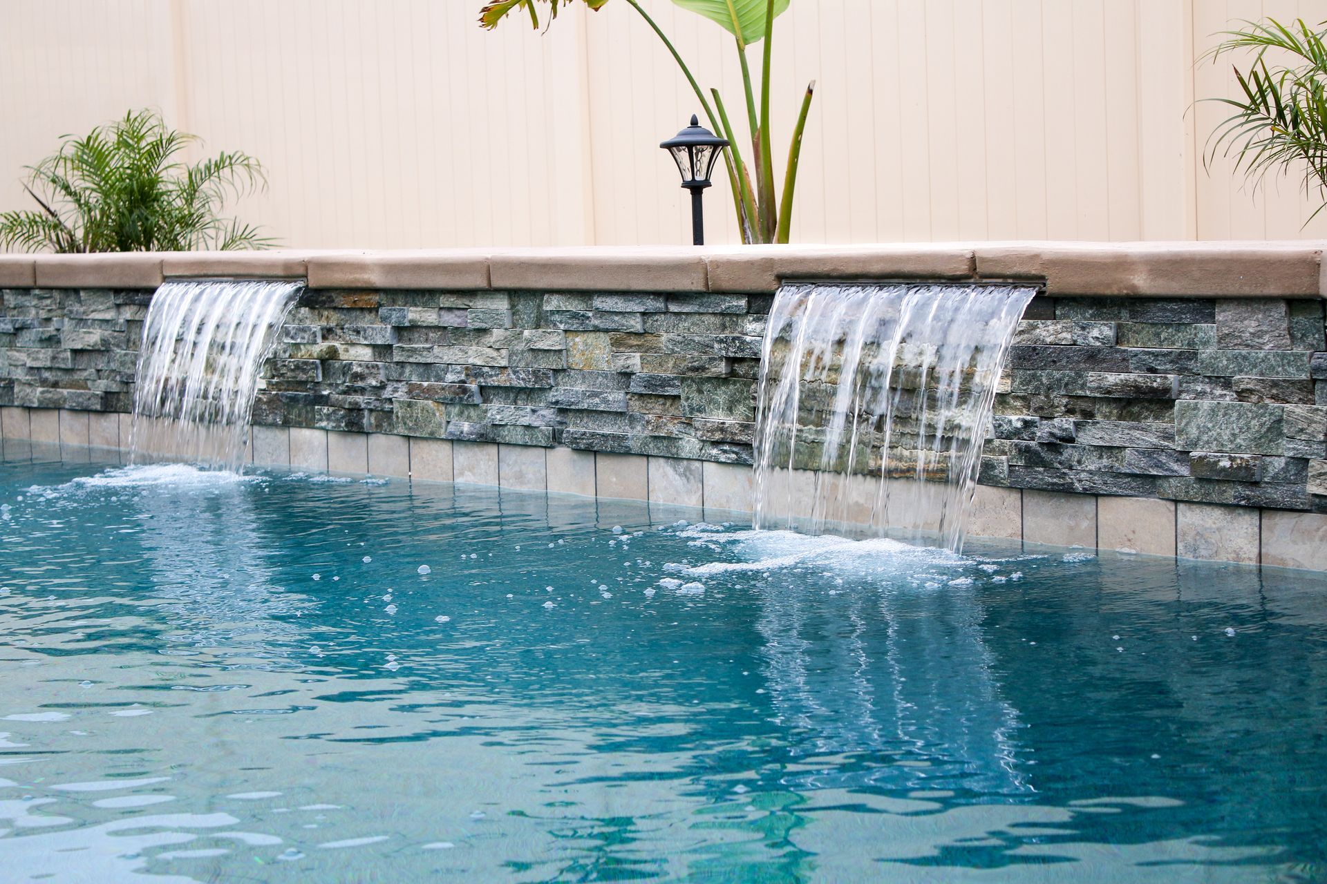 Two stone waterfalls pour water into a blue swimming pool in a backyard setting.