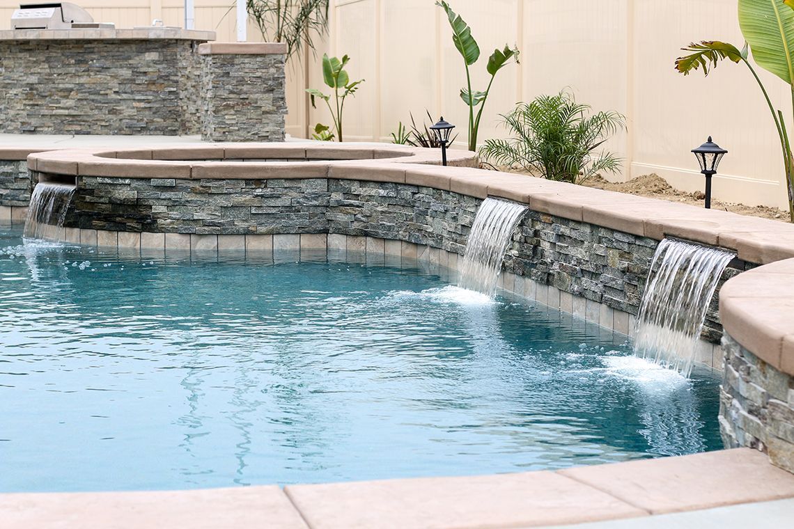 A blue swimming pool with a stone wall feature, tiered spa, and three small cascading waterfalls in a backyard.