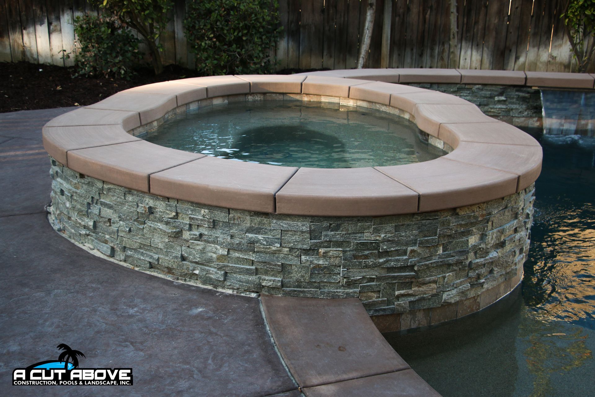 An elevated circular hot tub with stone-textured siding and tan coping stones, positioned next to a swimming pool.
