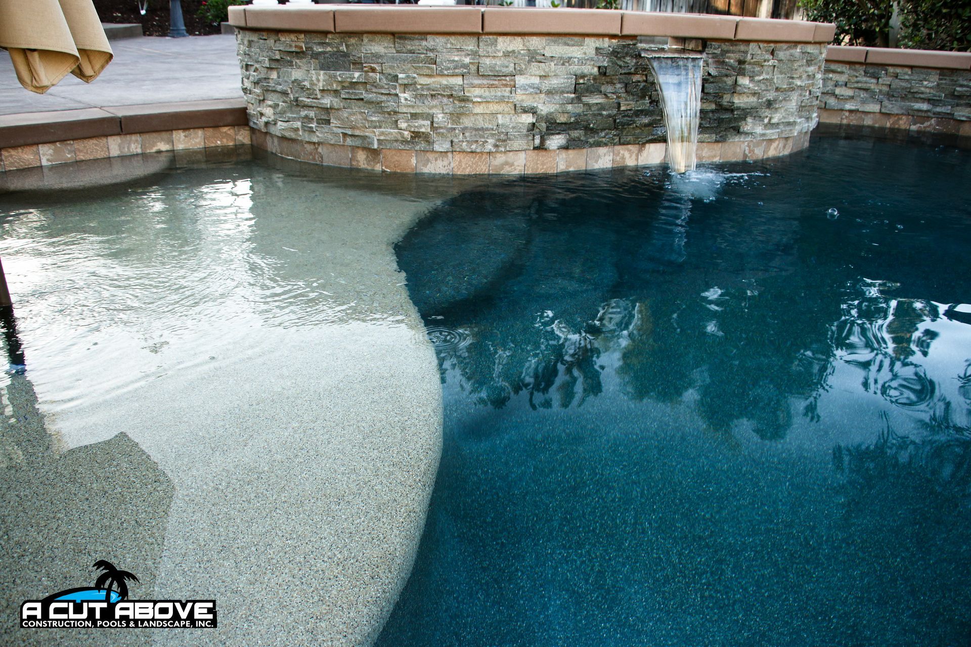 A swimming pool with a shallow tanning ledge, a deep blue main pool area, and a stone water feature pouring into it.
