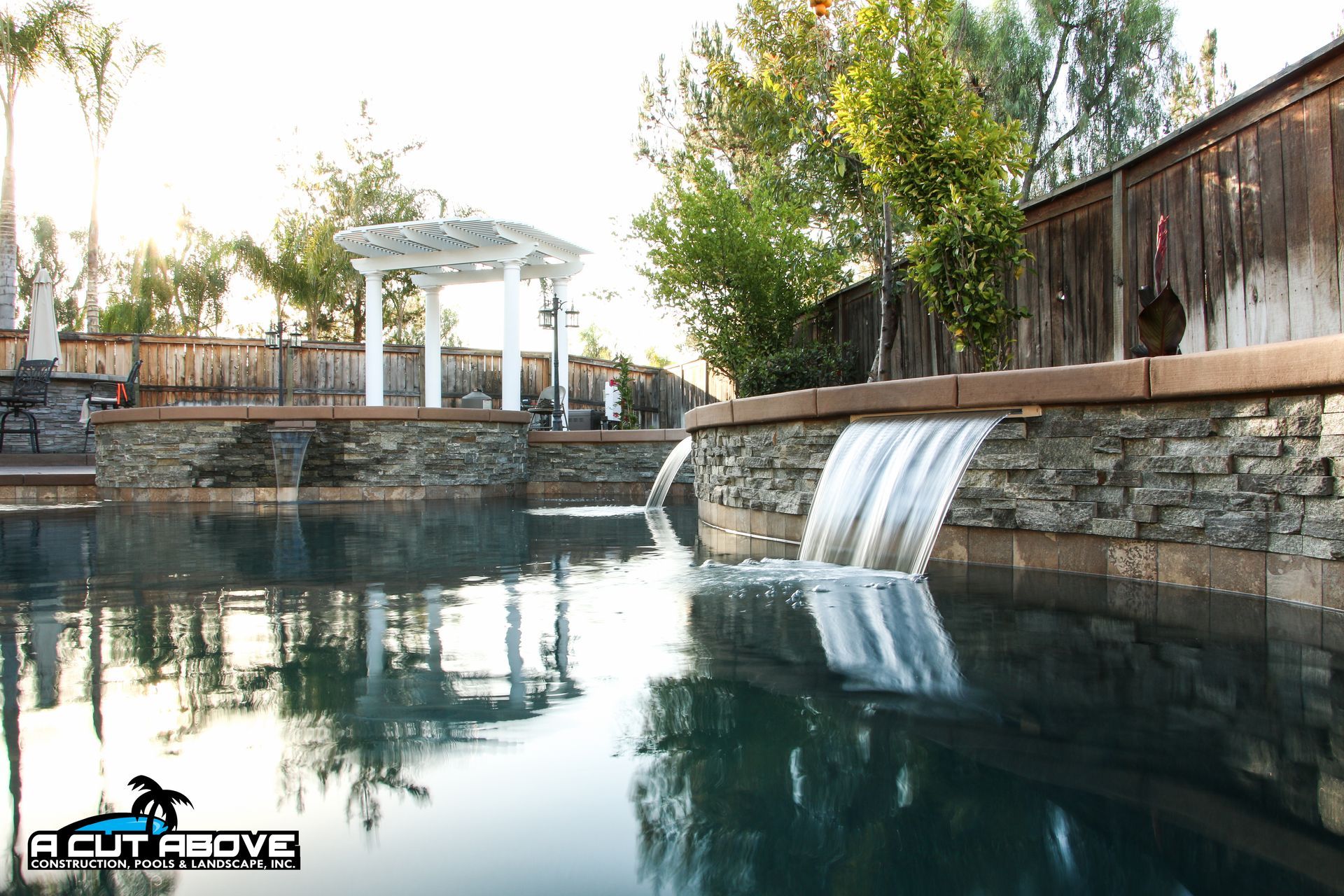 A backyard swimming pool with a stone wall, multiple water features, and a white pergola against a wooden fence.
