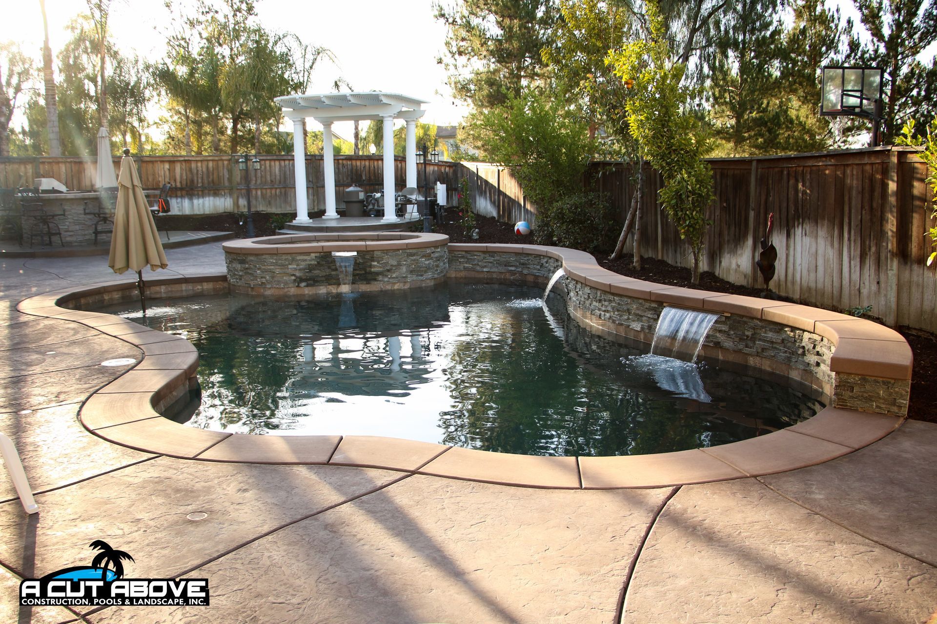 A backyard swimming pool with a stone-accented water feature, spa area, and white pergola on a stamped concrete patio.