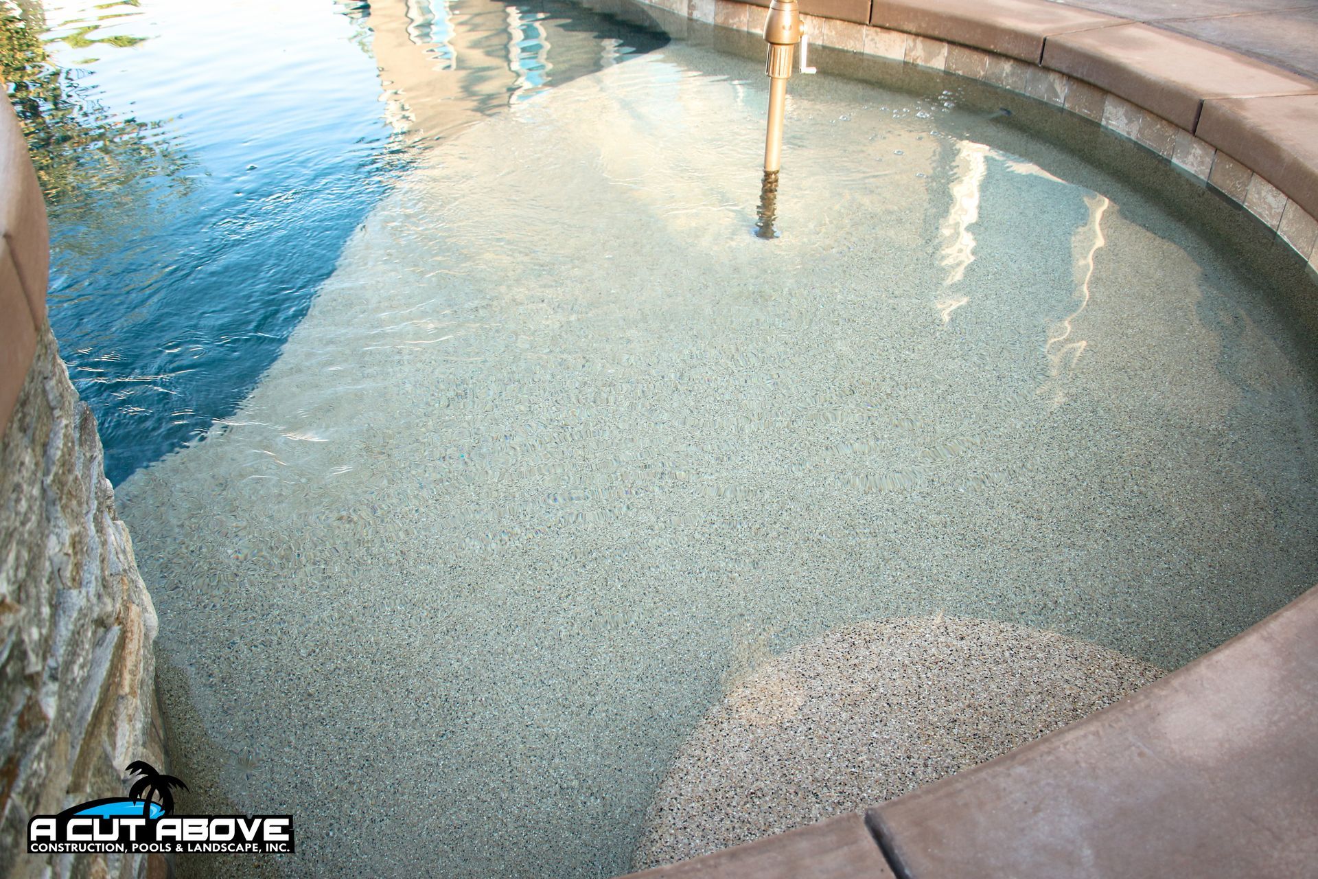 A curved, shallow sun shelf entrance to a swimming pool with light-colored pebble finish and stone coping.
