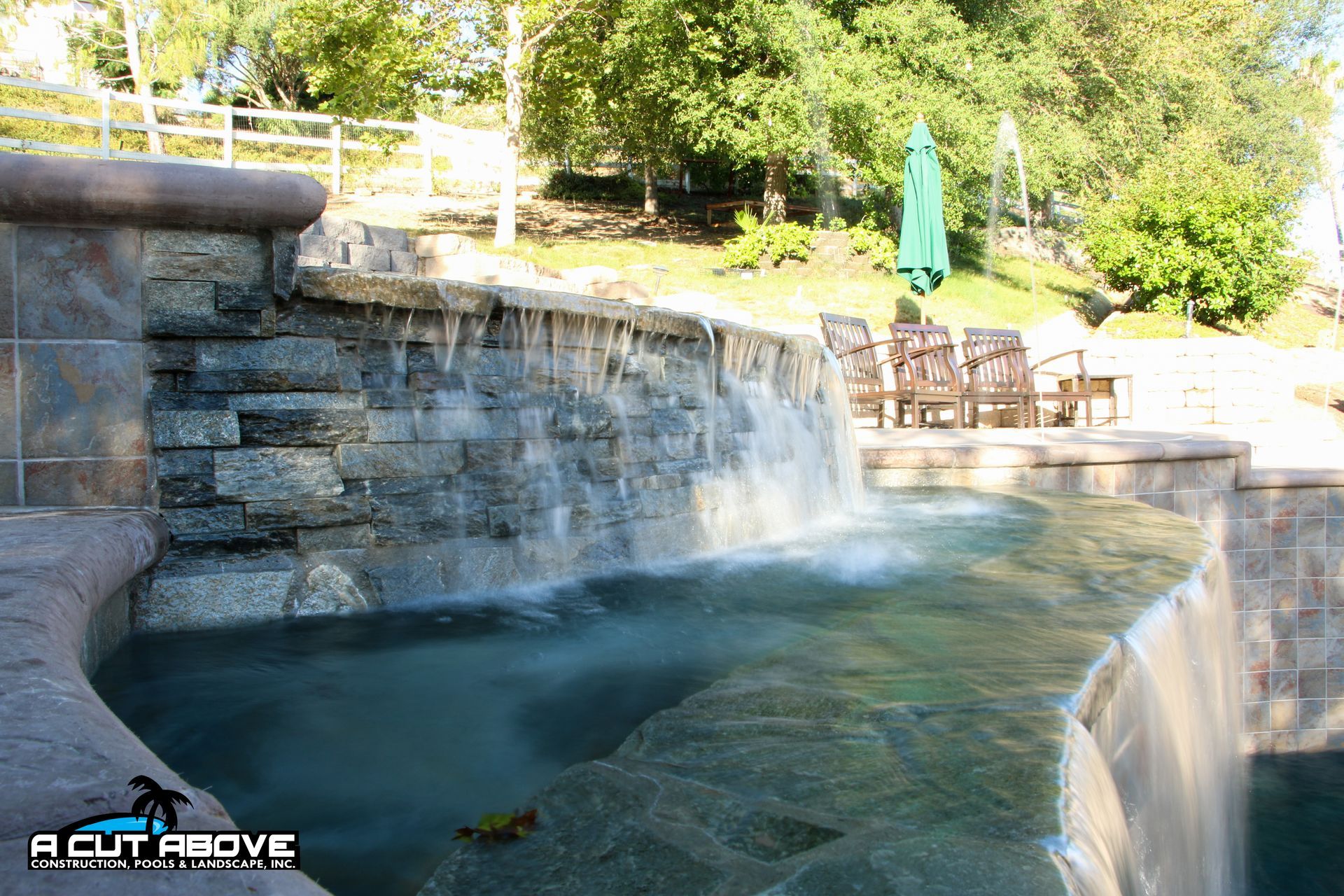 Stone waterfall feature with water flowing into a swimming pool, surrounded by trees and patio furniture.