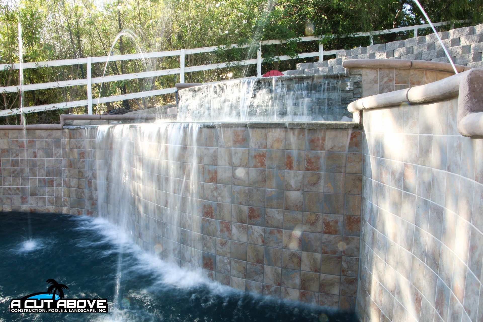 A stone tiered water feature spills into a pool, with a white fence and green trees in the background.