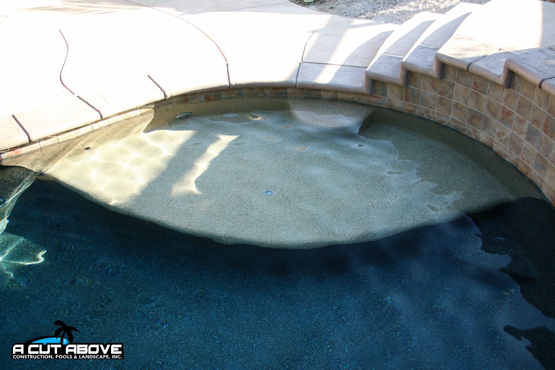 A curved pool ledge with built-in steps, featuring light-colored stone and dark water, adjacent to a decorative wall.